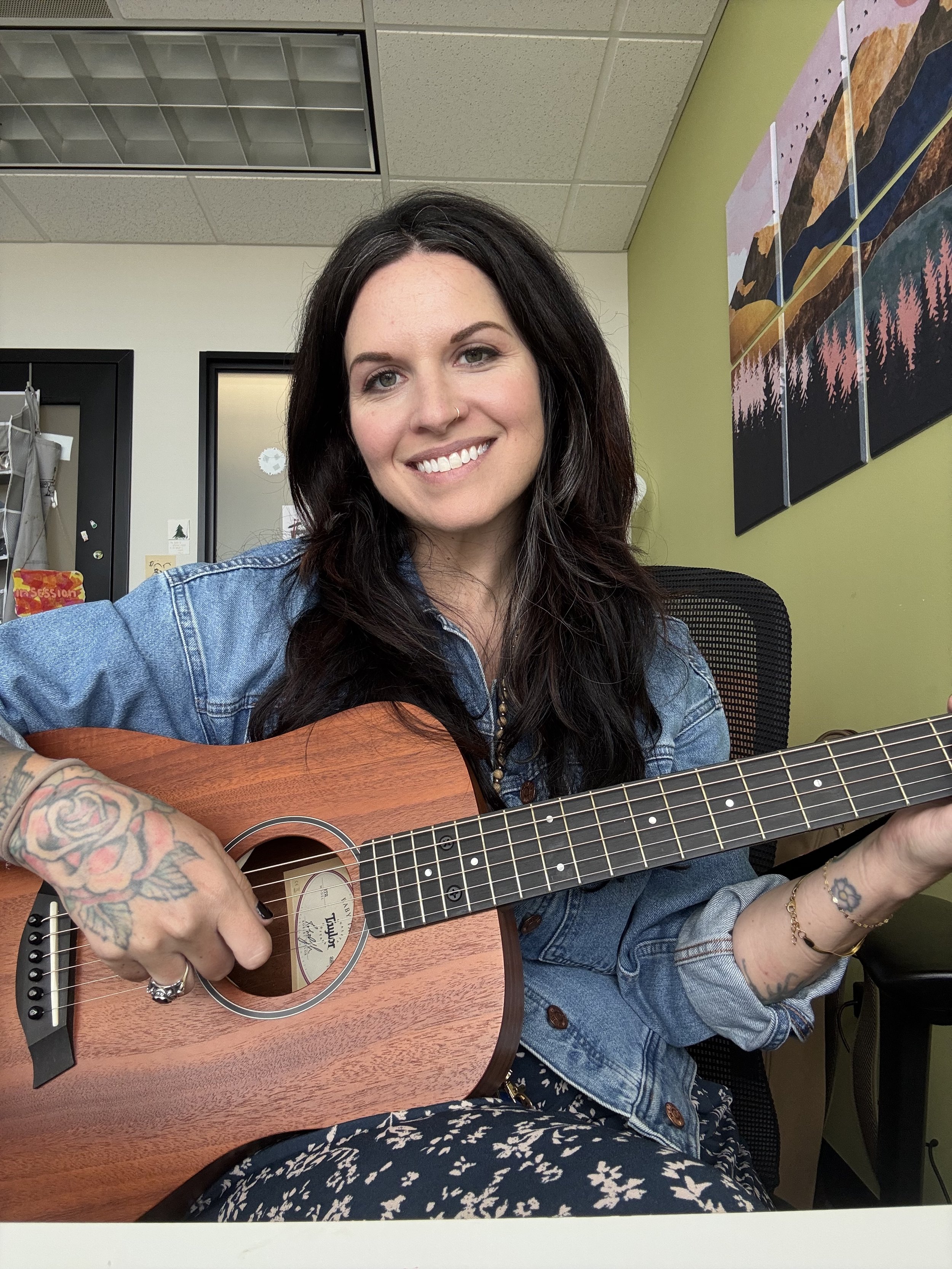 A woman with long dark hair wearing a denim jacket and floral dress playing an acoustic guitar, smiling, sitting in an office or studio with a landscape painting on the wall.