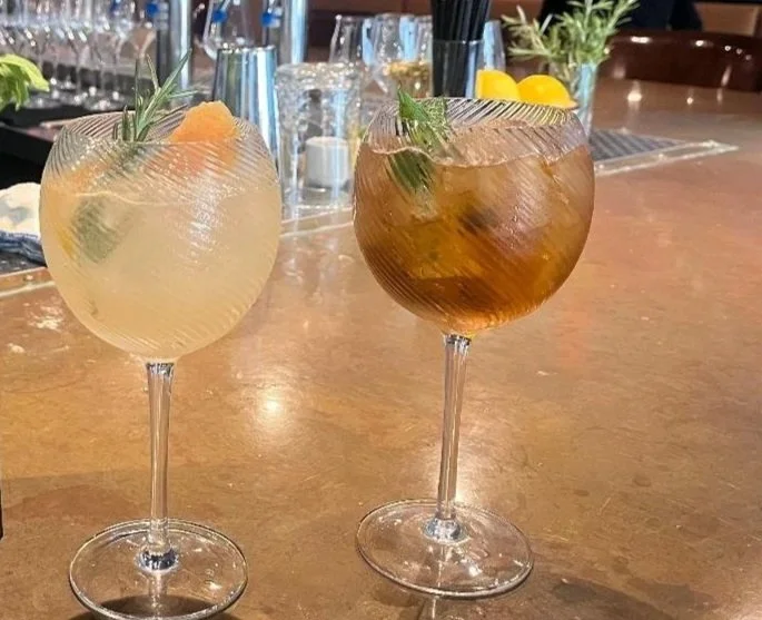 Two large cocktail glasses with hearty drinks, one golden and one dark, garnished with mint and citrus slices, on a bar or restaurant counter.