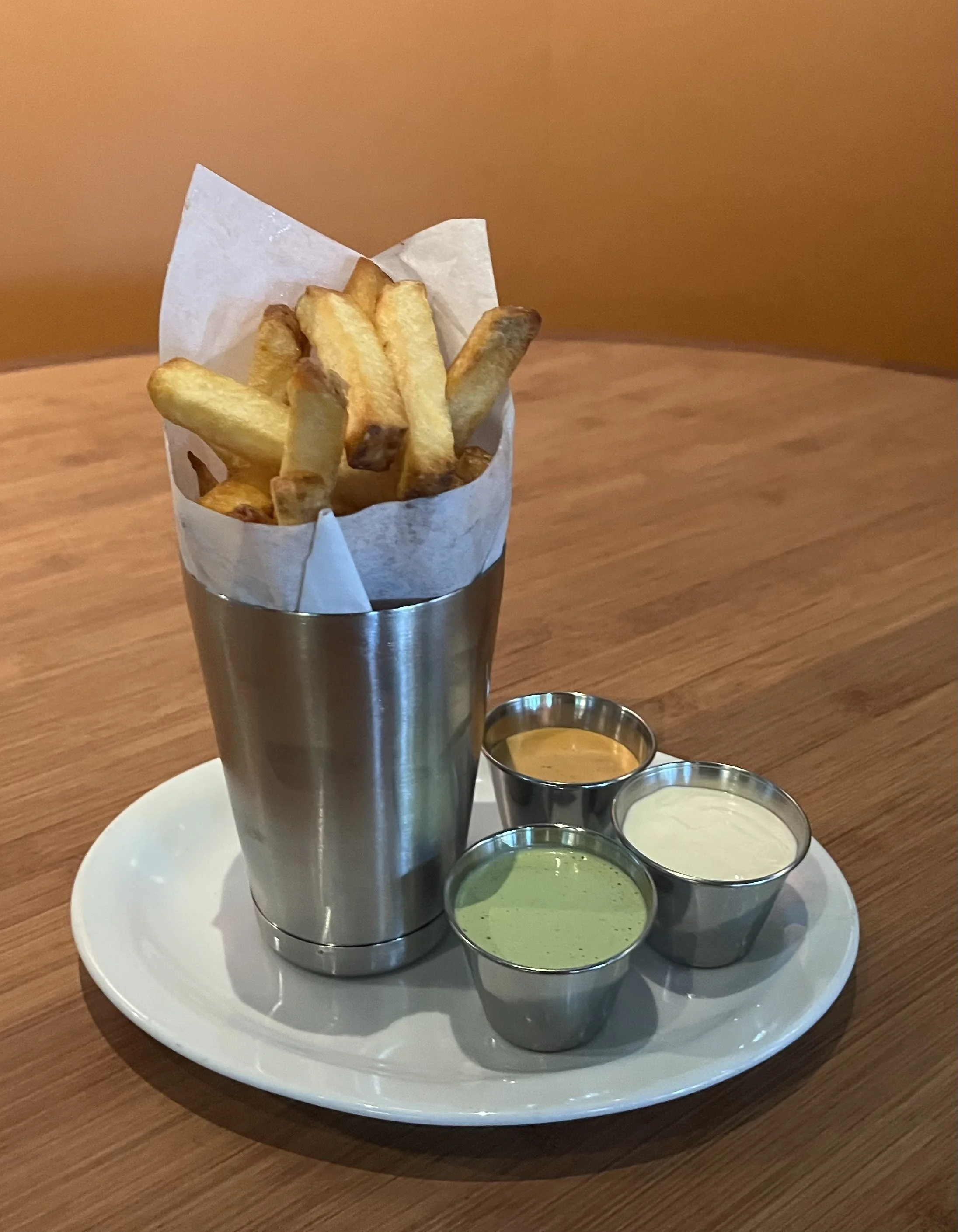 A metal cup filled with French fries with four different dipping sauces on a white oval plate.