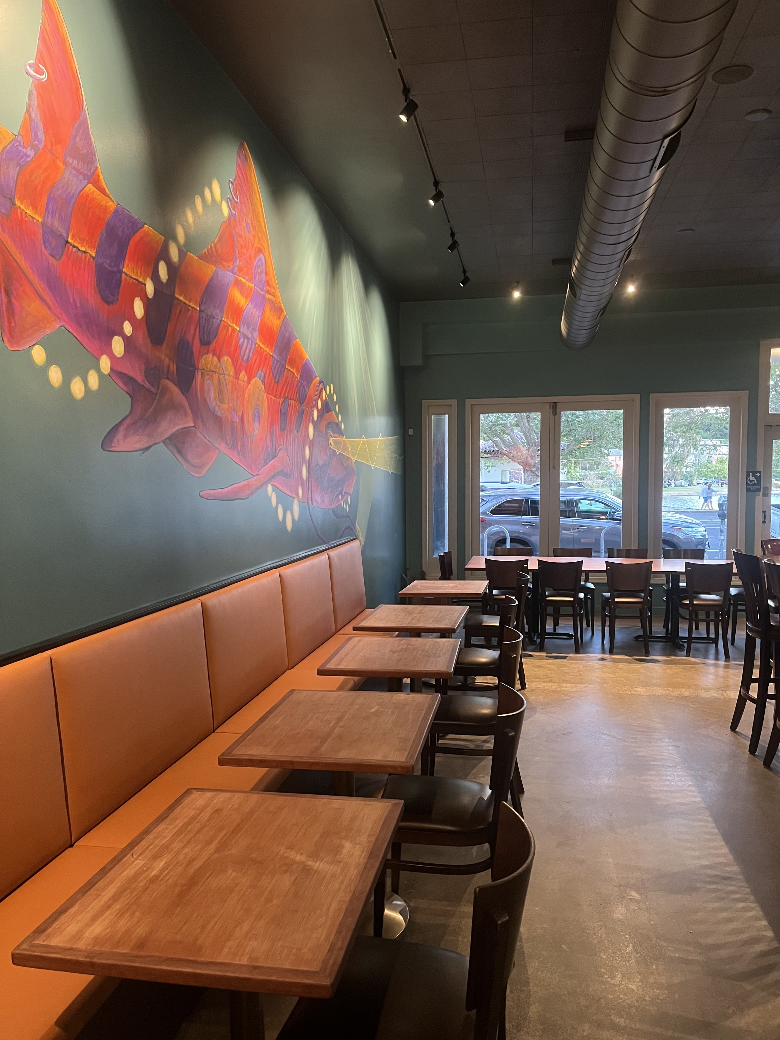 Interior of a cafe with a green wall, a large colorful fish mural, orange seating, wooden tables, and black chairs. Large windows show the parking lot outside, and a person is walking on the sidewalk.