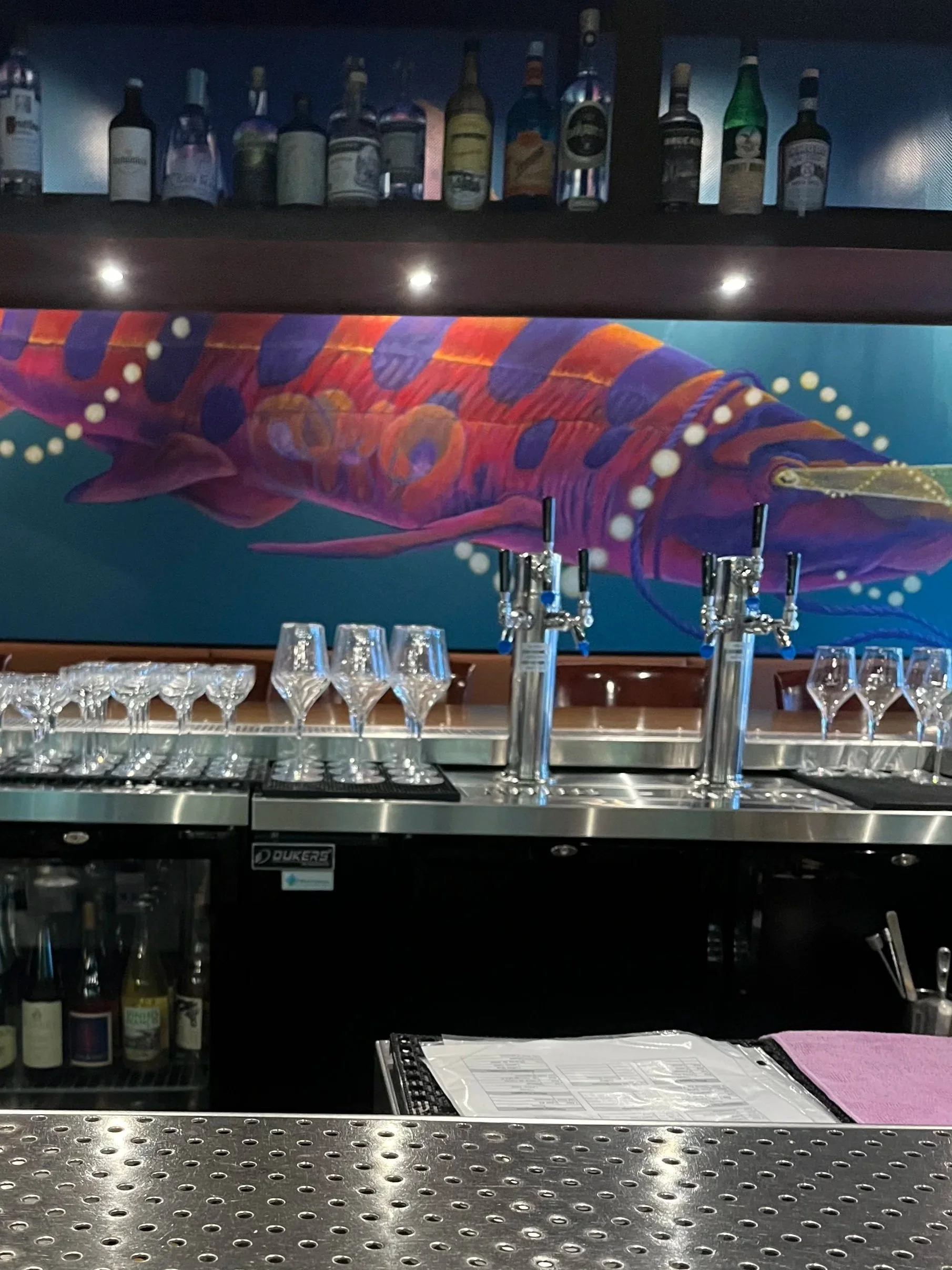 Bar with a colorful fish mural, bar counter with glasses, and liquor bottles on shelf