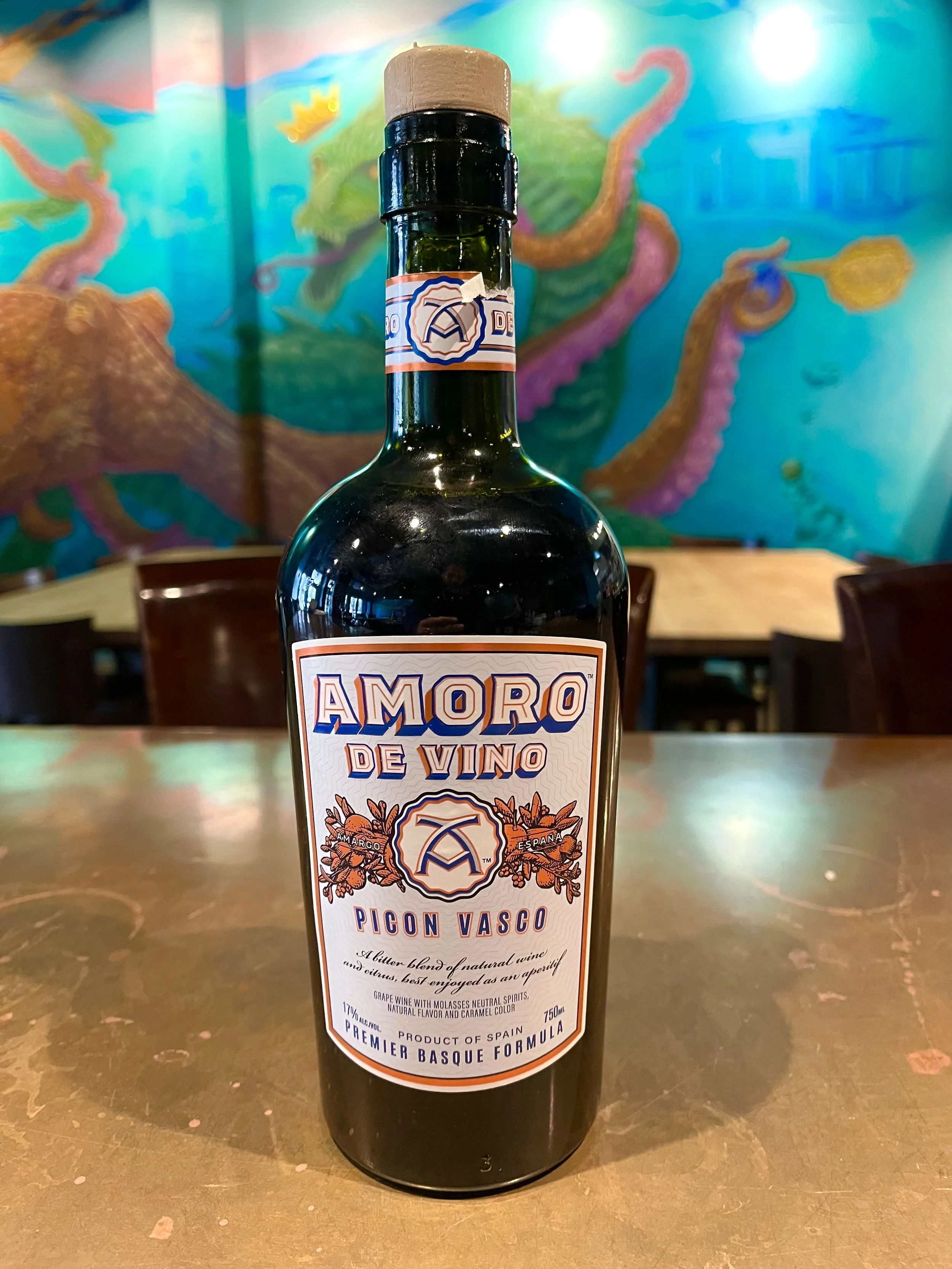A wine bottle labeled 'Amoro de Vino Picon Vasco' on a table, with a colorful mural of a dragon in the background.
