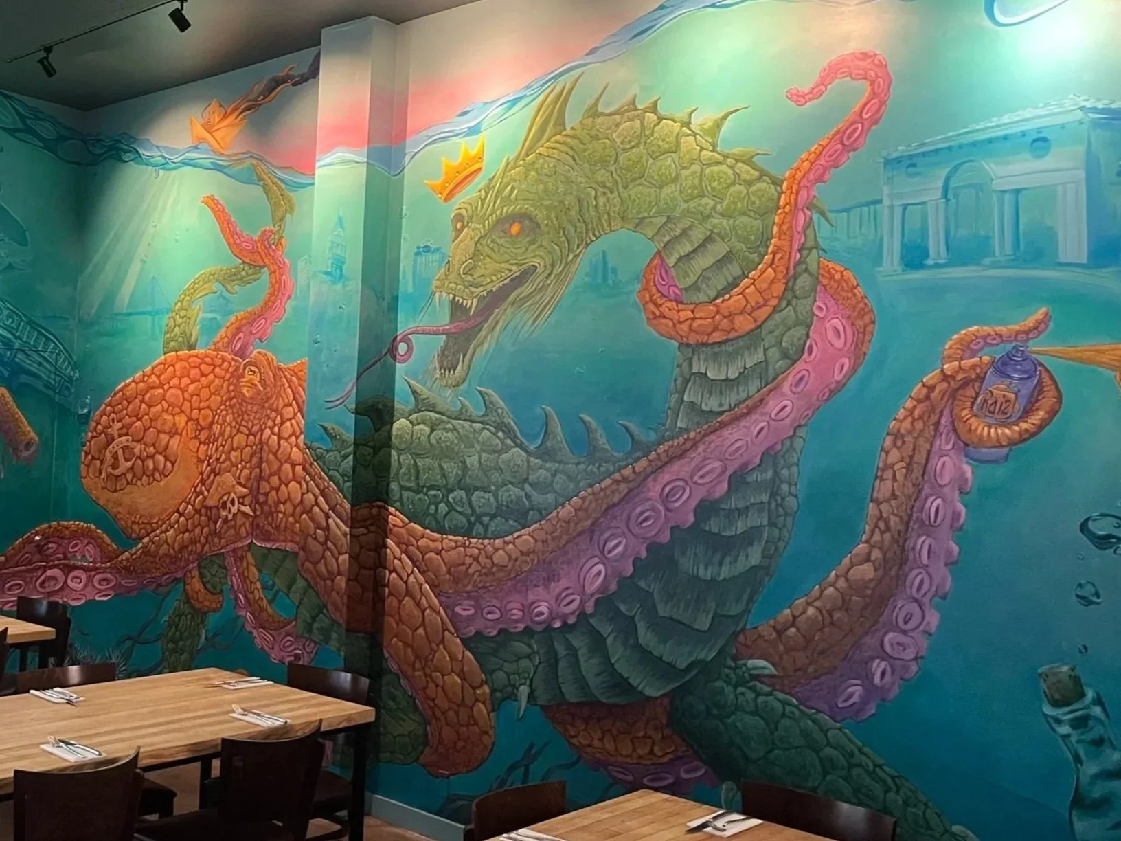 Colorful mural of a giant sea monster with a dragon-like head and an octopus body, holding a spray paint can, in a restaurant setting.
