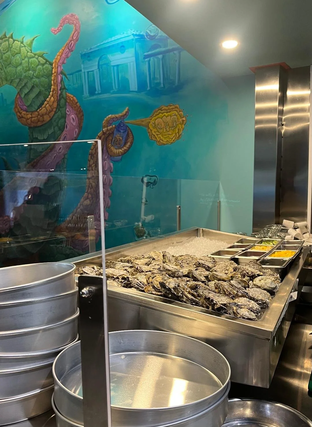 A seafood display with oysters on ice at a restaurant, with a colorful underwater mural featuring a sea creature and a building in the background.