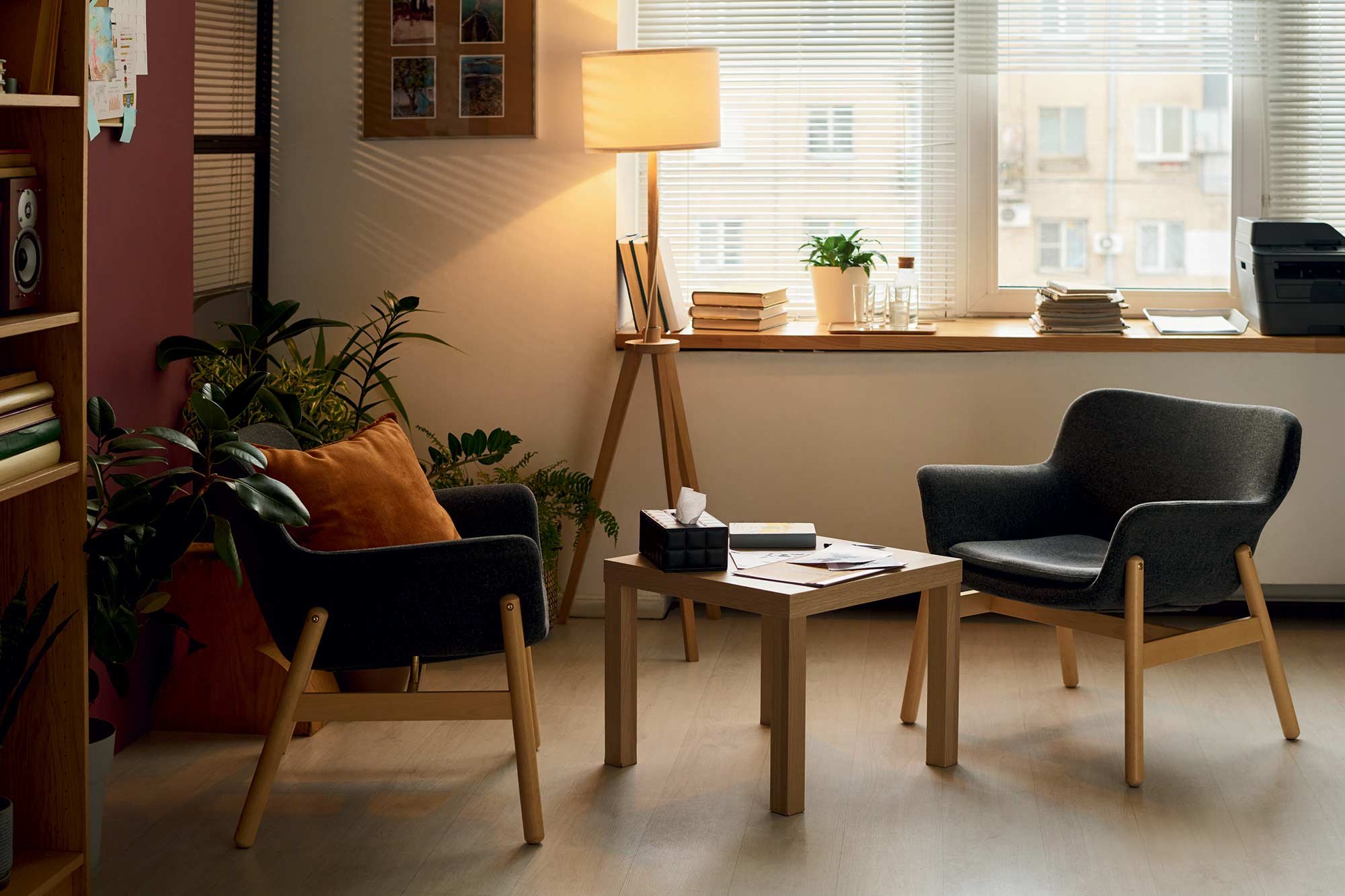 A cozy office corner with two black armchairs, a small wooden coffee table with papers and a tissue box, a wooden desk with stacks of papers, a potted plant, and a window with blinds letting in natural light.