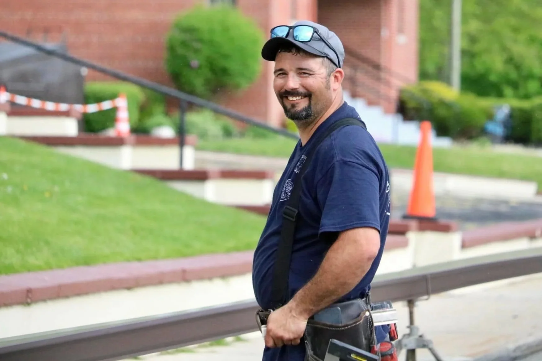 A smiling man wearing a cap, sunglasses, and a work uniform, standing outdoors near a metal railing with construction cones in the background.