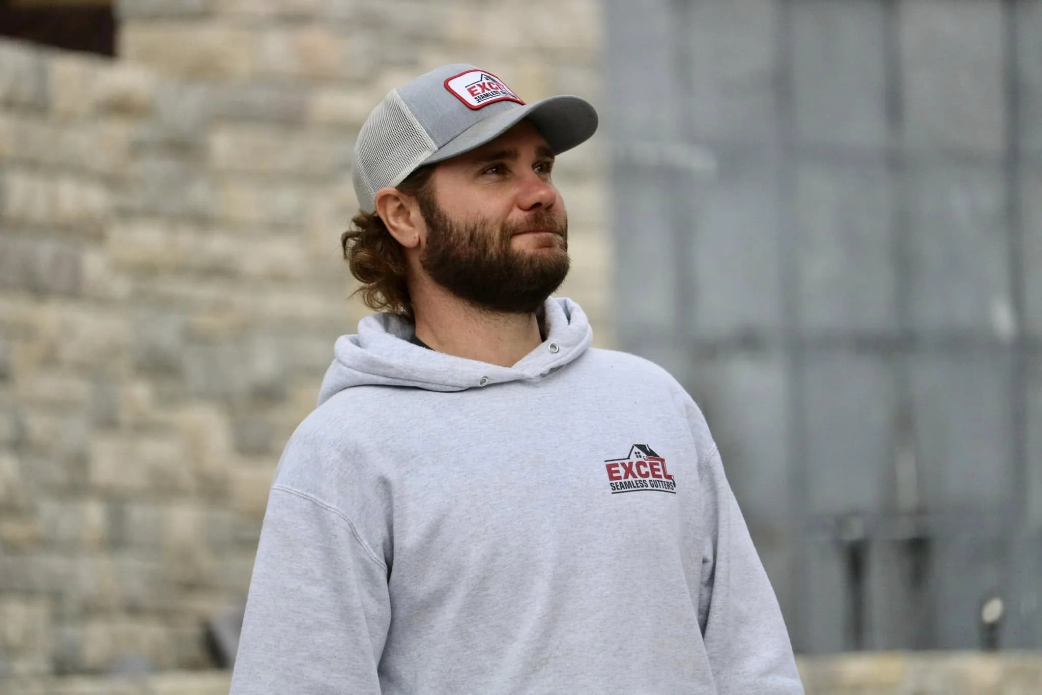 A man with a beard and long hair wearing a gray baseball cap and a light gray hoodie with an 'Excel Seamless Gutters' logo on it, standing outdoors near a stone wall.