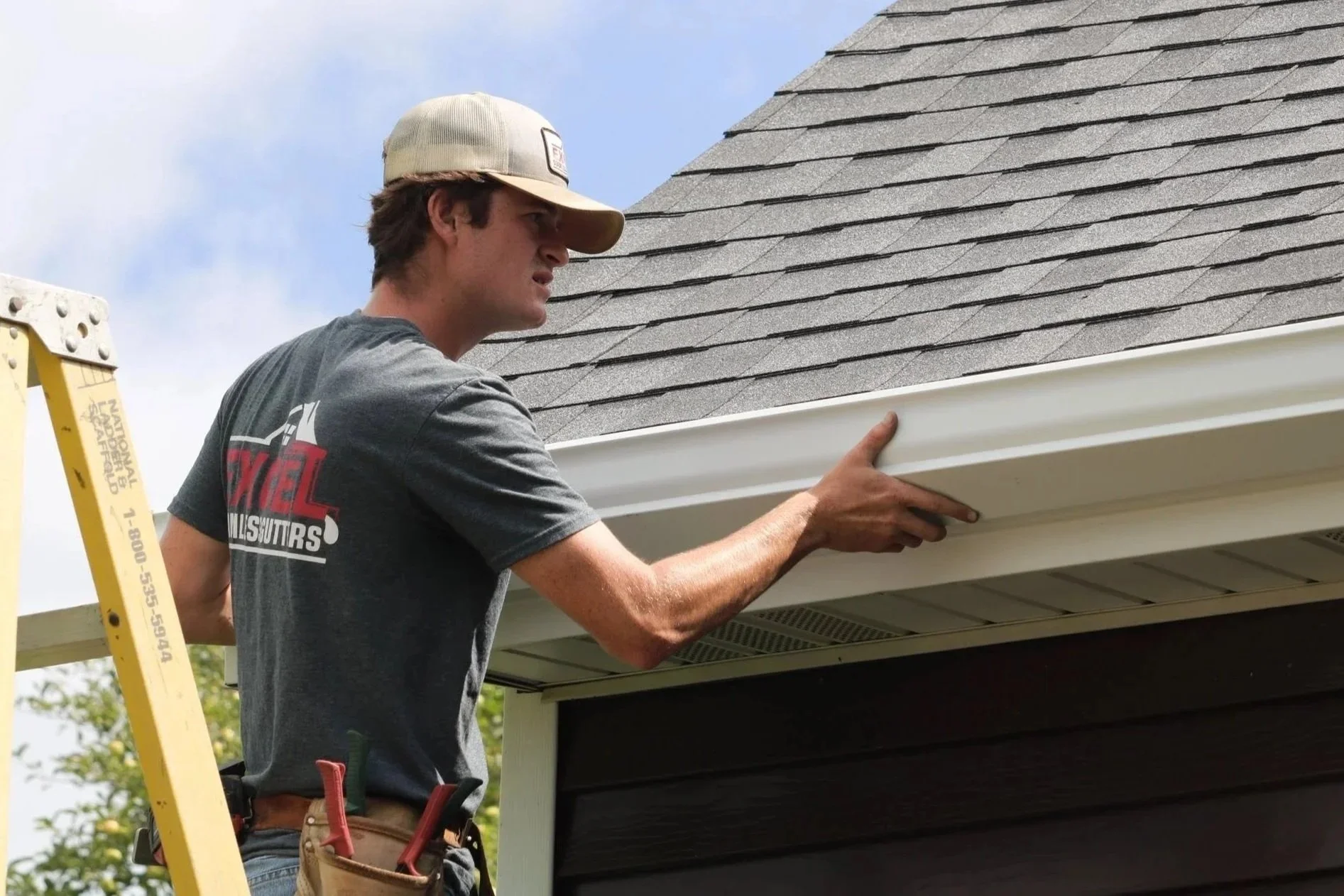 A man wearing a baseball cap and a gray T-shirt with a logo, standing on a yellow ladder, installing or inspecting white trim on a house's exterior corner during the daytime. The house has gray shingles on the roof, and there are trees and a partly cloudy sky in the background.