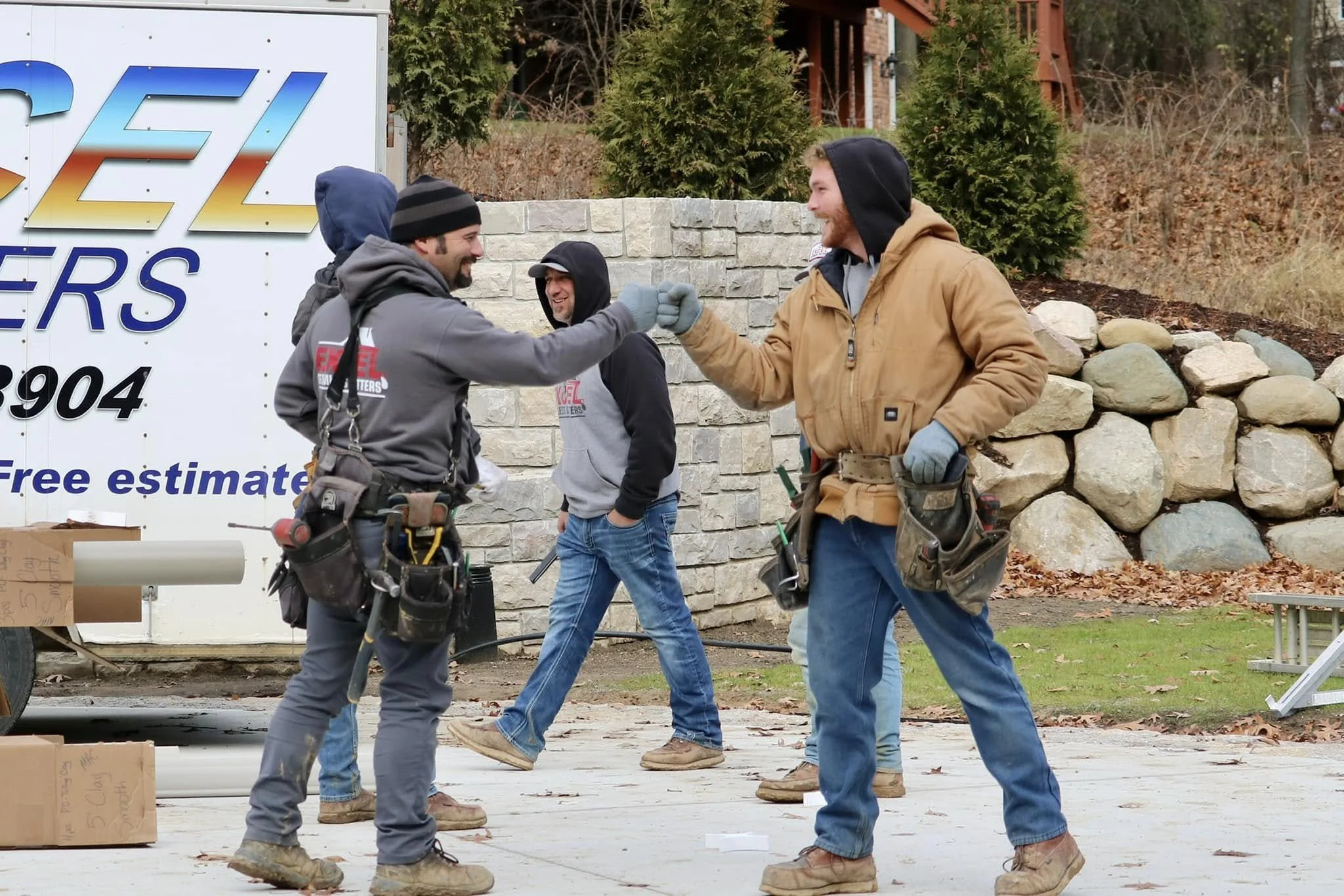 Two men are smiling and fist-bumping outdoors, wearing work clothes and gloves, with a third man standing behind them, in front of a stone wall and green trees.