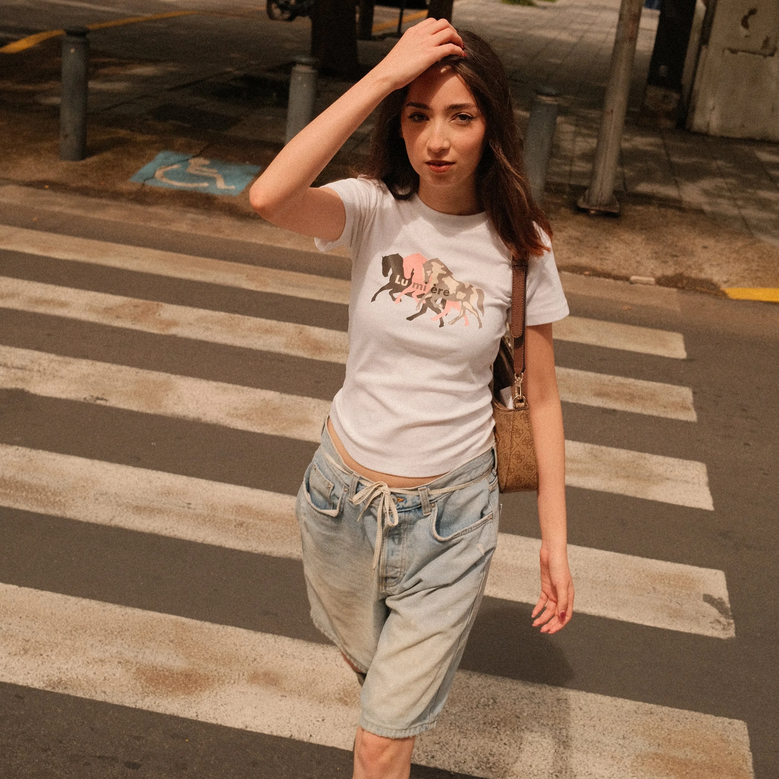 Young woman with long dark hair wearing a white t-shirt with horse silhouettes and the words 'Lu miére,' paired with light blue shorts, standing outdoors at night on a crosswalk near parking lot and sidewalk.