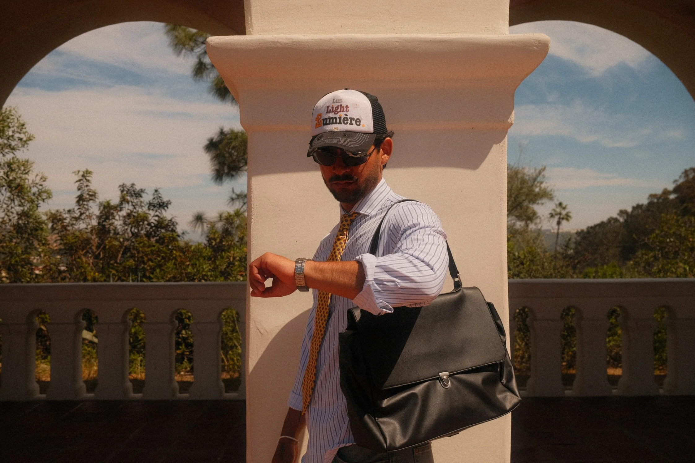 A man wearing sunglasses, a cap that reads 'Luz Light La Lumière', a striped shirt, and a yellow tie, looking at his watch, with a black bag over his shoulder, standing outdoors against a white wall with decorative columns and green trees in the background.