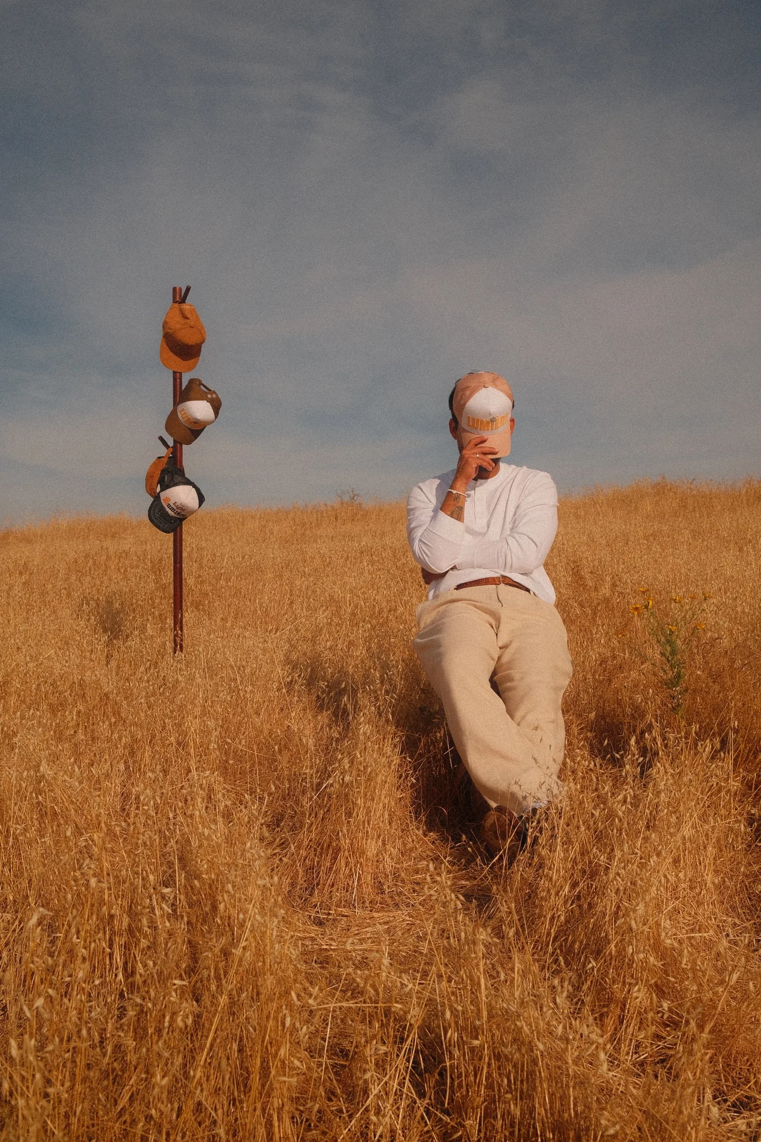 A person sitting in a golden wheat field with baseball caps hanging on a wooden stick nearby, wearing a white shirt and beige pants, holding a cap in front of their face under a partly cloudy sky.