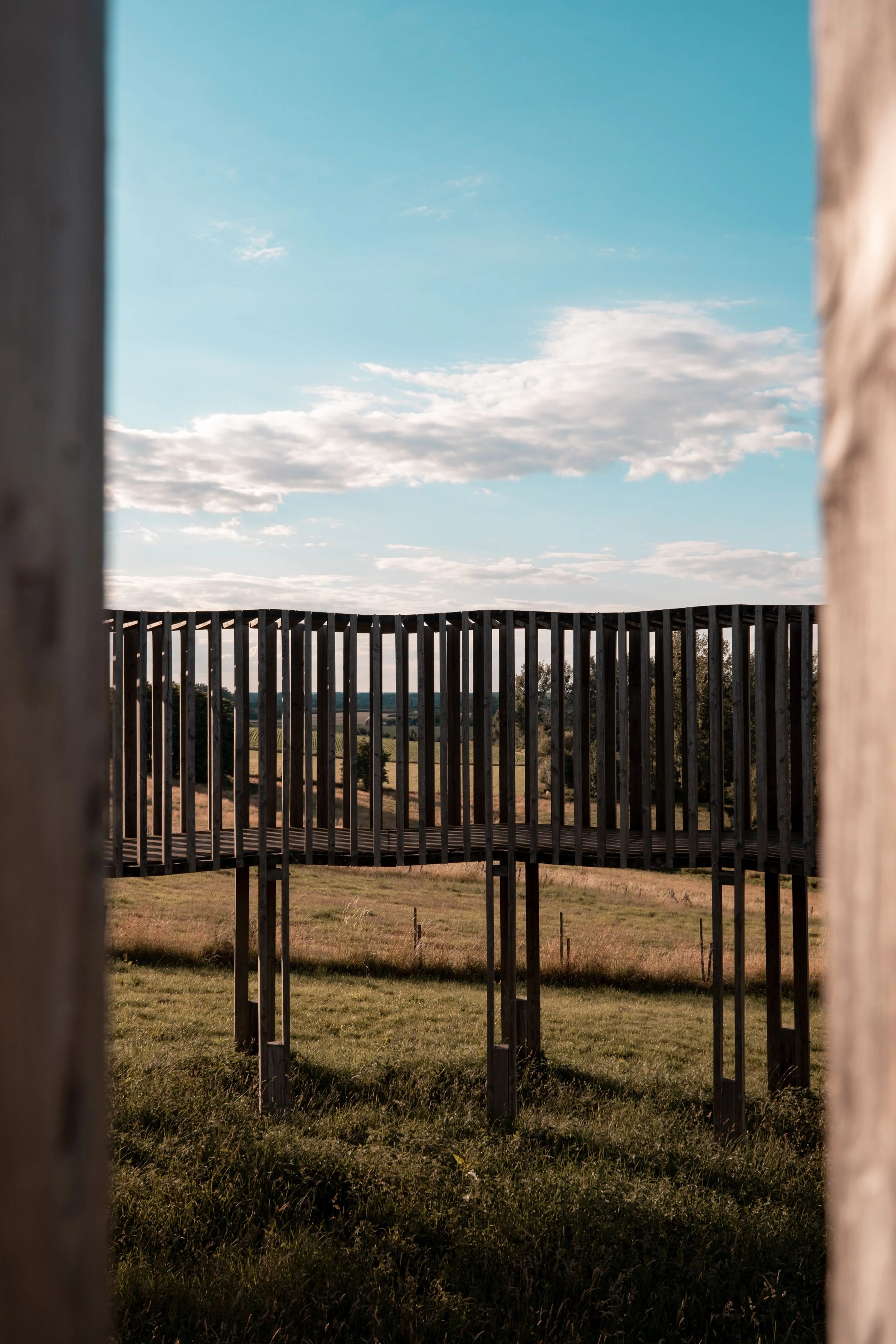 A view through a wooden frame or structure showing a grassy field and a wooden fence with a curved design, under a sky with scattered clouds.