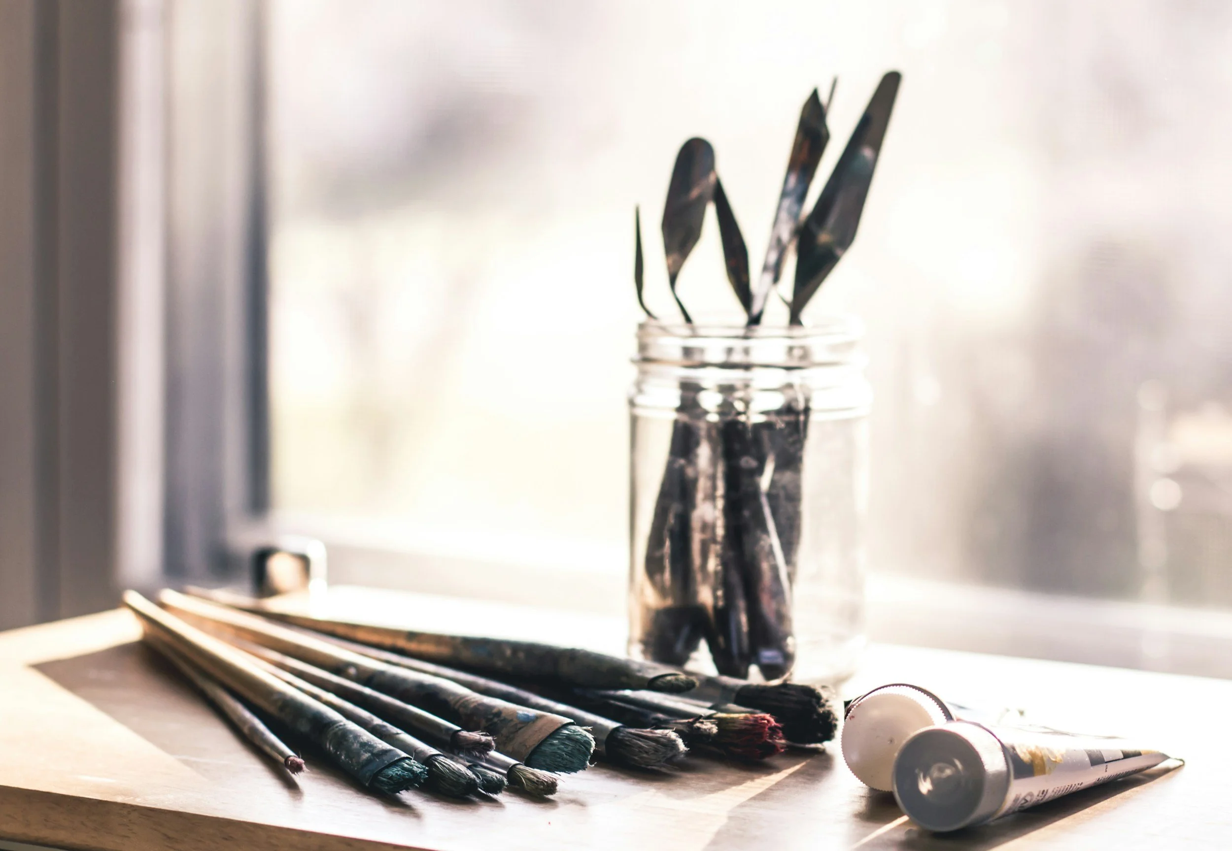 A collection of used paintbrushes with paint on the bristles, a jar, and some tubes of paint on a desk or table, with a window in the background.