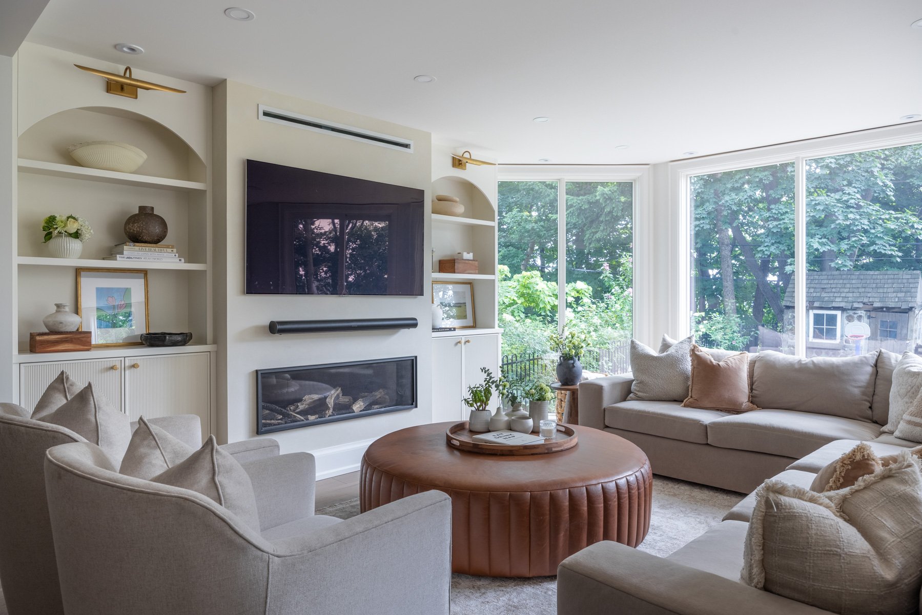 Bright living room with large windows overlooking green trees. Features a beige sectional sofa with multiple pillows, a round brown leather ottoman, built-in shelves with decorative objects, a flat-screen TV, and a fireplace.