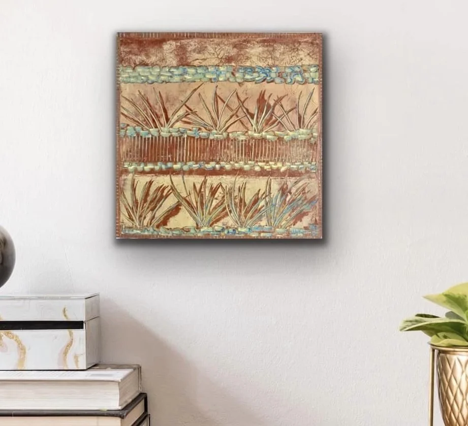 Decorative square wall art with abstract earthy tones and plant patterns, hung on a white wall above a white vanity with a marble geometric pattern, and next to a potted green plant with large leaves.