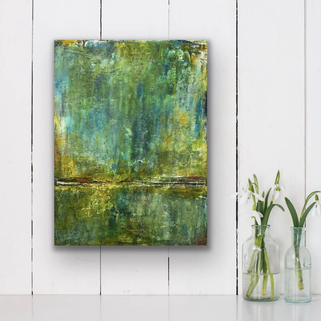 Abstract green and yellow painting hanging on a white wooden wall next to two glass vases with white flowers.
