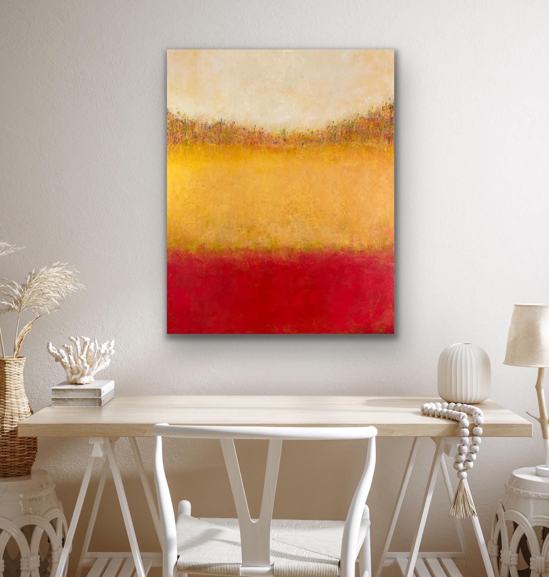 A room with a white wall, featuring a large abstract painting with warm colors including yellow, orange, red, and cream. There is a wooden table with white chairs, and decorative items such as white coral, a woven basket with pampas grass, and two va