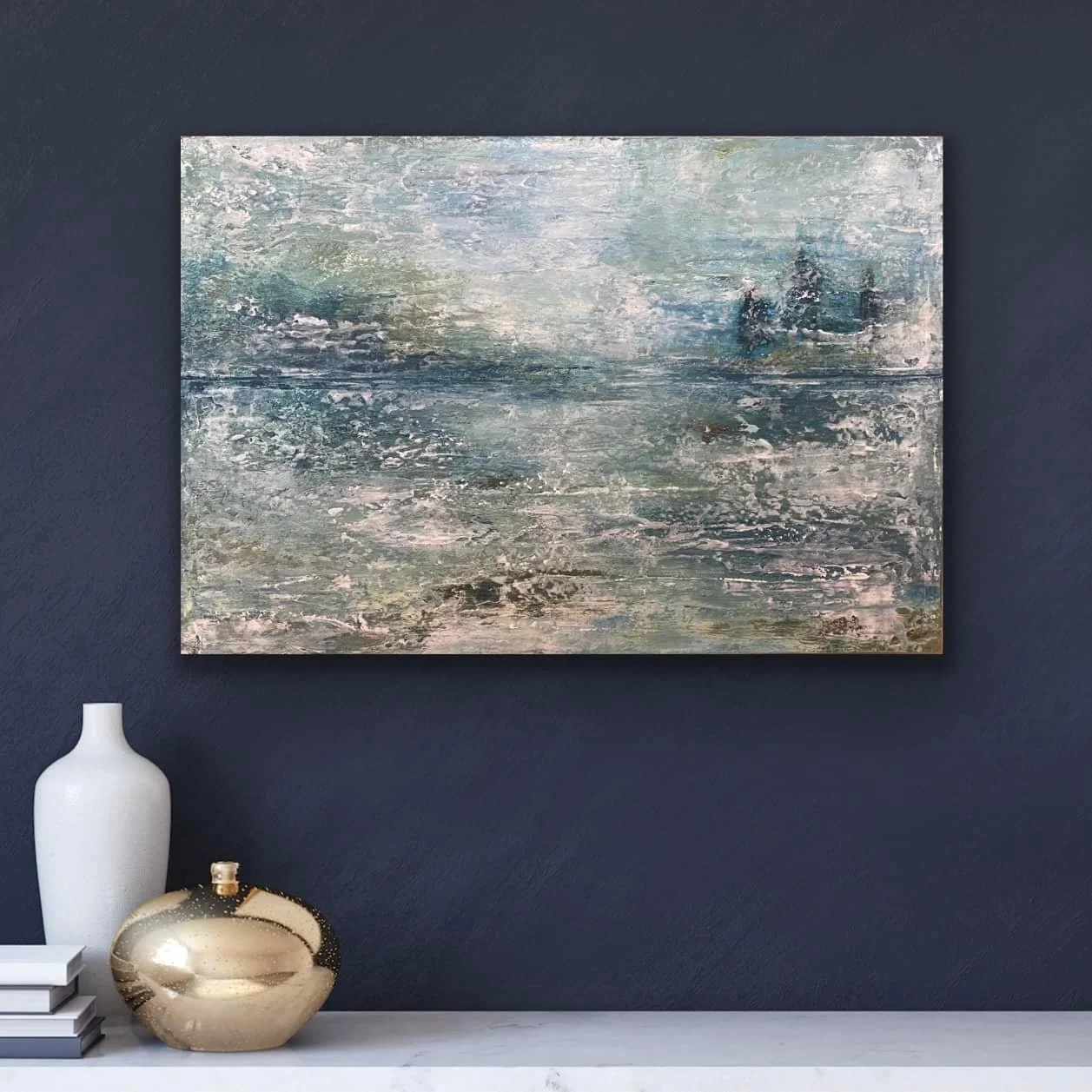Abstract landscape painting with shades of blue, green, and gray hanging on a dark wall above a white shelf. The shelf holds a tall white vase, a metallic gold decorative object, and a stack of books.
