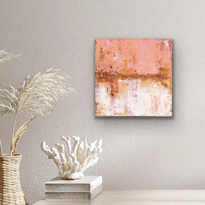 Abstract pink, orange, and white painting hanging on a gray wall, with a wicker vase holding dried pampas grass and a white coral sculpture on top of two stacked books on a white surface.