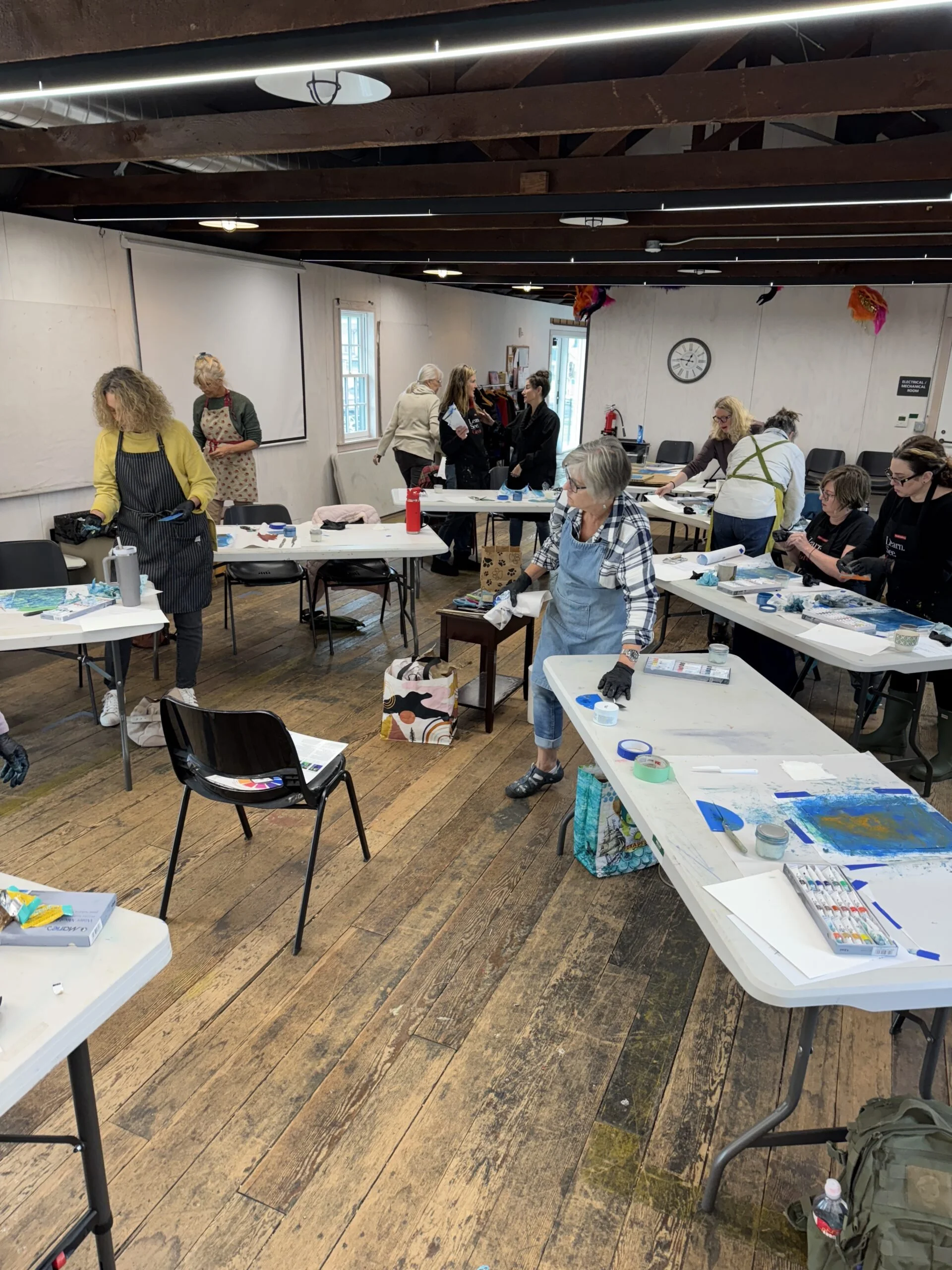 A group of women participating in an arts and crafts workshop in a spacious room with wooden floors and exposed beams on the ceiling. Tables are covered with art supplies, and some women are working on projects while others are standing and chatting.