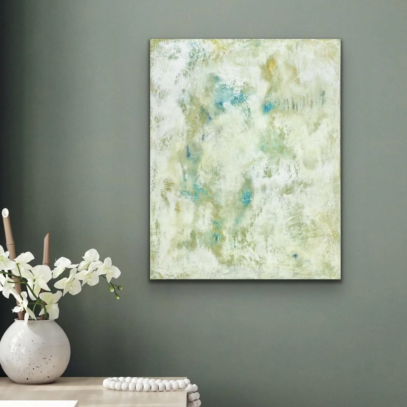 A decorative interior wall with a large abstract painting, a white vase with white orchids, and a pearl-like bead garland on a beige surface.