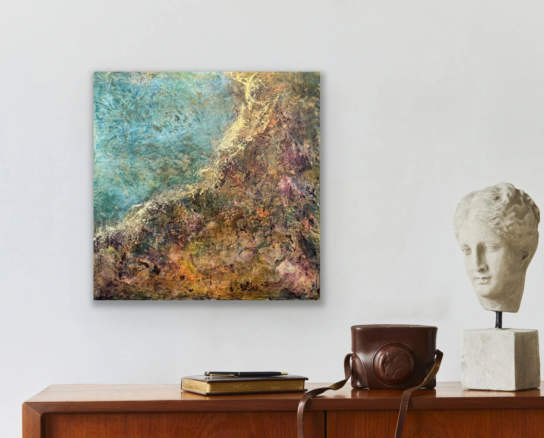 A close-up of a wooden surface with an art piece and decorative items arranged on it. The art piece is a colorful textured painting with blue, green, yellow, purple, and brown tones. To the right, there is a bust sculpture of a head, likely made of p