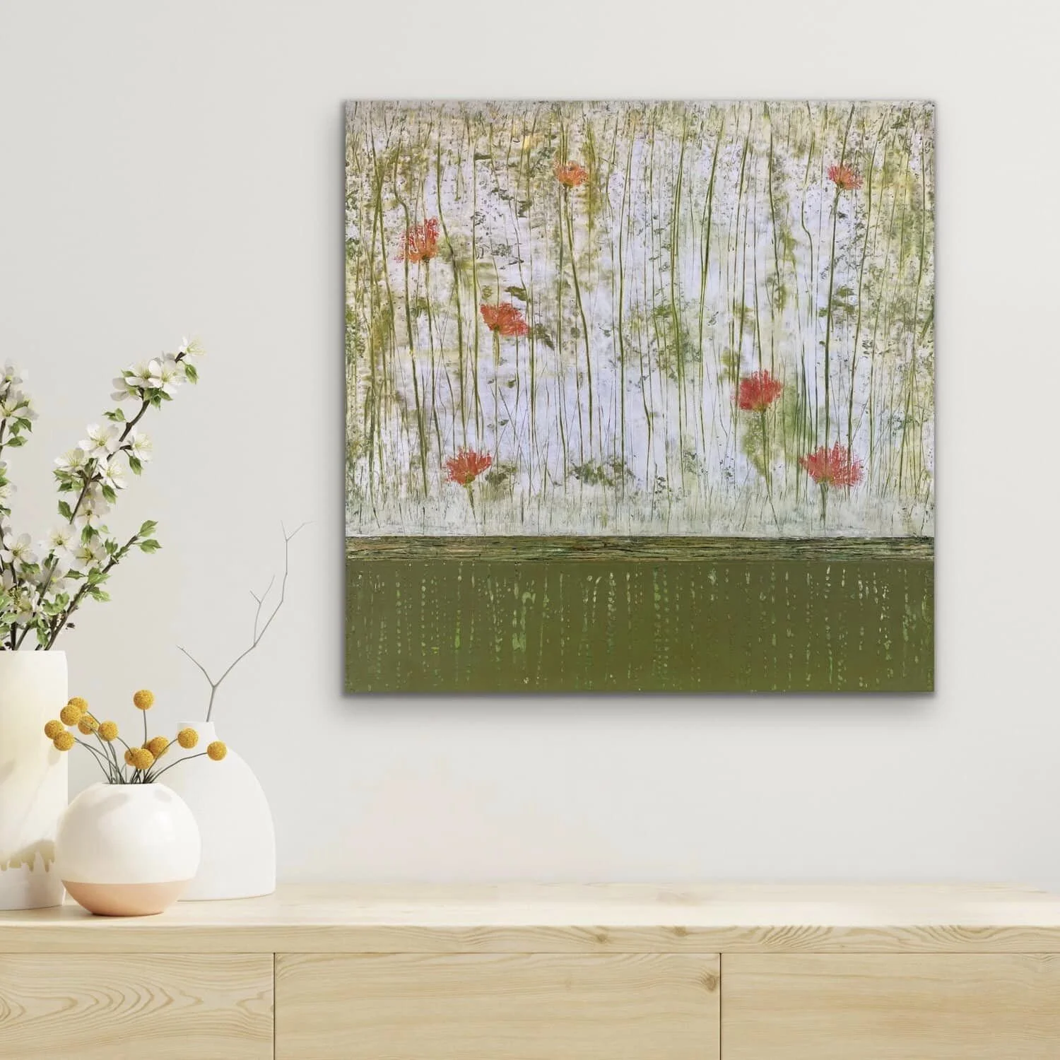 A modern interior wall with a large abstract painting featuring a pond scene with lily pads and reeds, above a light wooden sideboard. On the left, white and beige vases with flowering branches and yellow flowers.