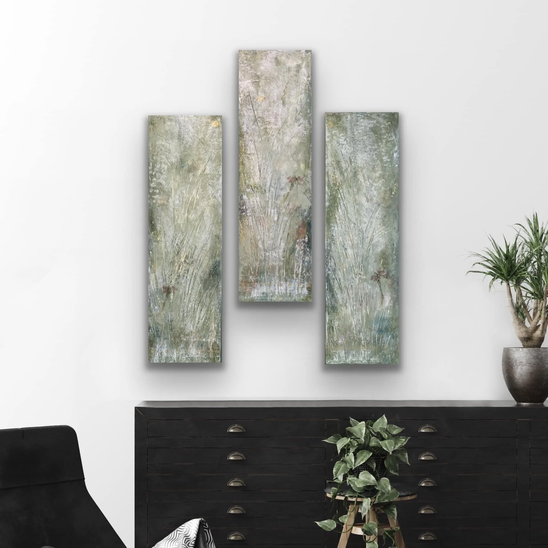 Black wooden dresser with six drawers, silver handles, a potted green ivy plant on the right, and three abstract landscape paintings hanging above. The paintings depict grassy fields, trees, and sky in soft pastel colors.