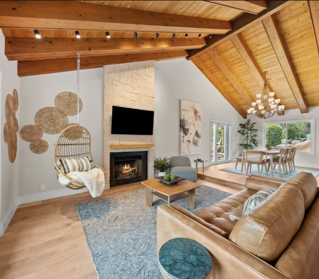 Living room with wooden ceiling, fireplace, mounted TV, hanging wicker chair, tan leather sofa, coffee table, gray armchair, textured wall art, green potted plant, dining table with chairs, large windows, and chandelier.