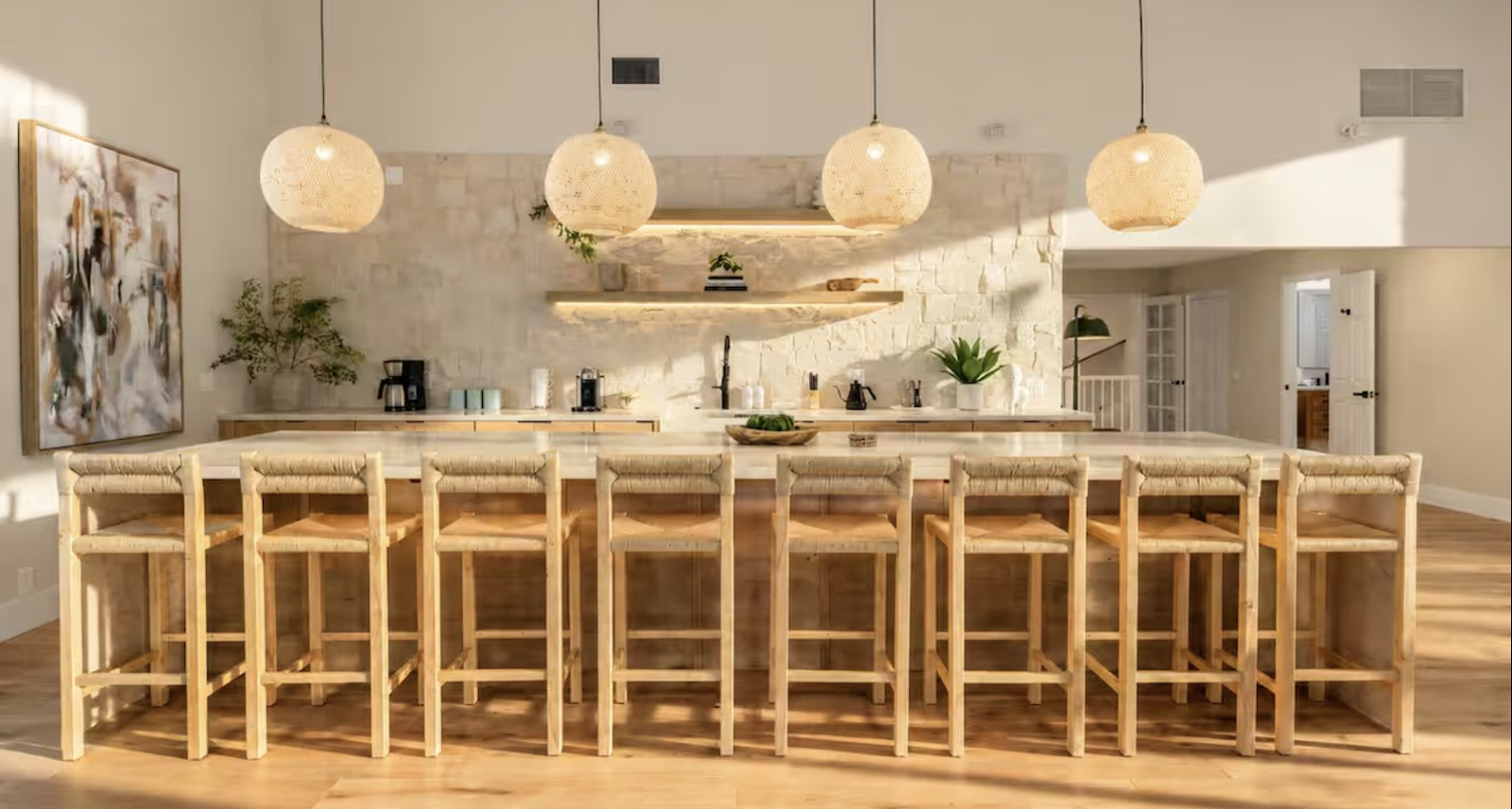 Modern kitchen with a long island counter and ten wooden barstools, pendant lights, white brick wall, green plants, and open doors in the background.
