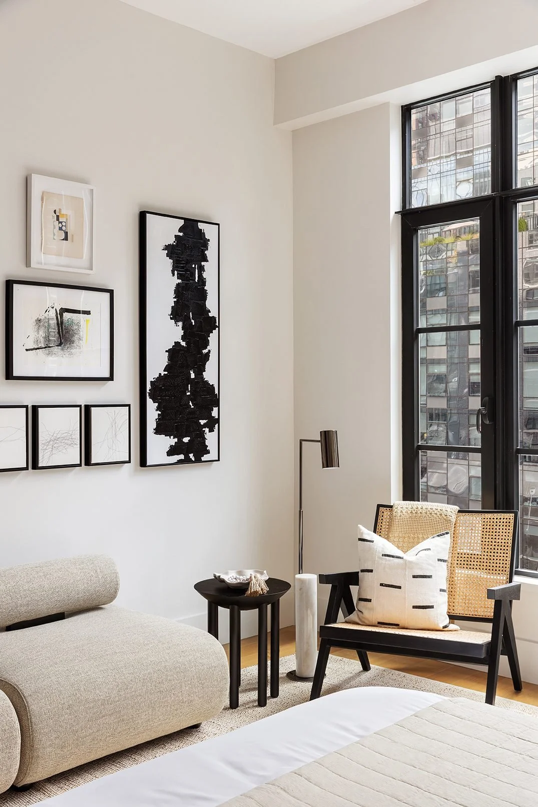 Photo of art direction for 517 West 29th - Modern bedroom corner with a beige sofa, black side table, black framed abstract art on white wall, black framed artwork, cane chair with a white pillow, tall floor lamp, and large windows showing cityscape.