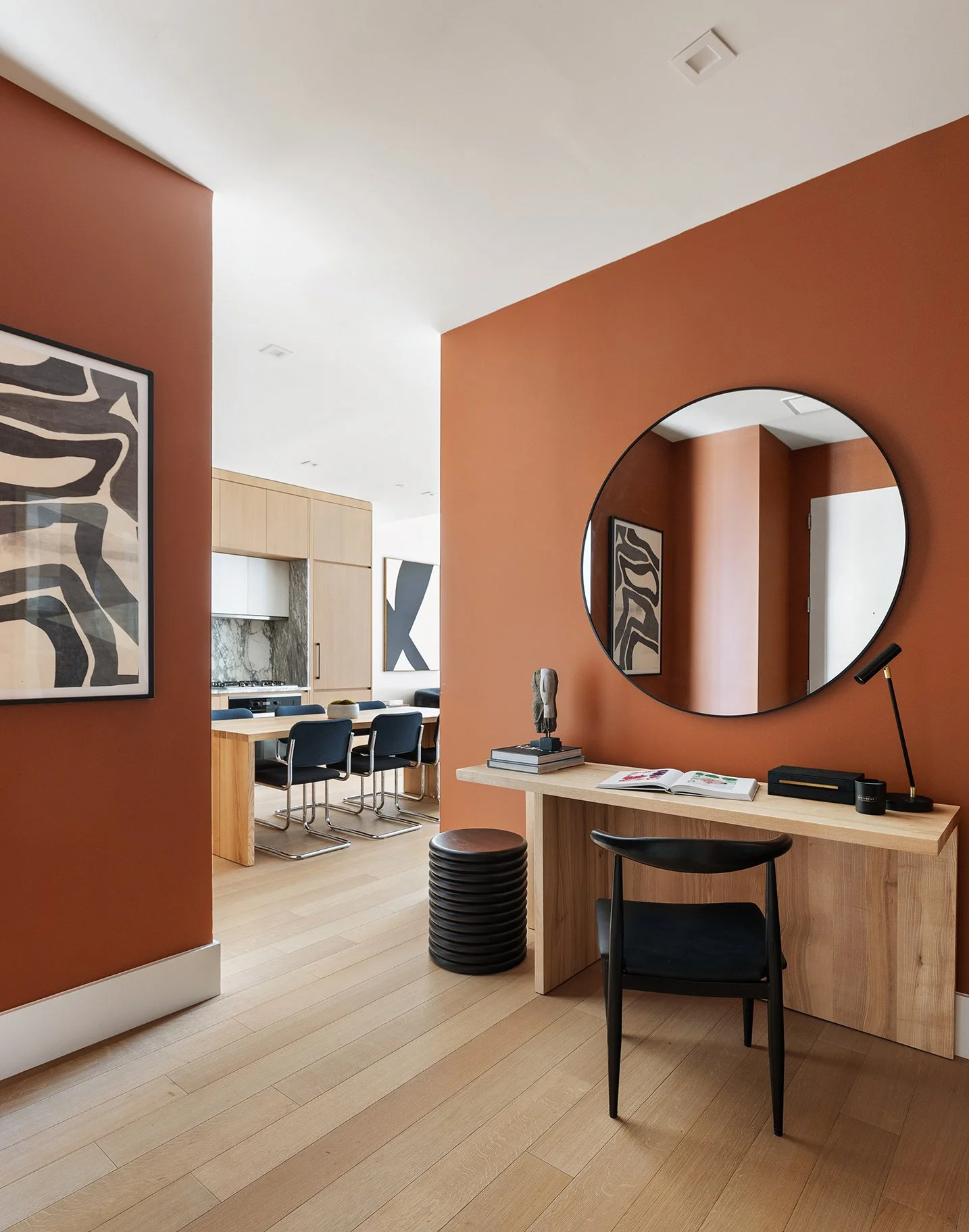 Photo of art direction for 517 West 29th - Modern dining and workspace area with an orange accent wall, a large round mirror, artwork, a wooden desk with a black chair, and a open-concept kitchen with a wooden table and black chairs.