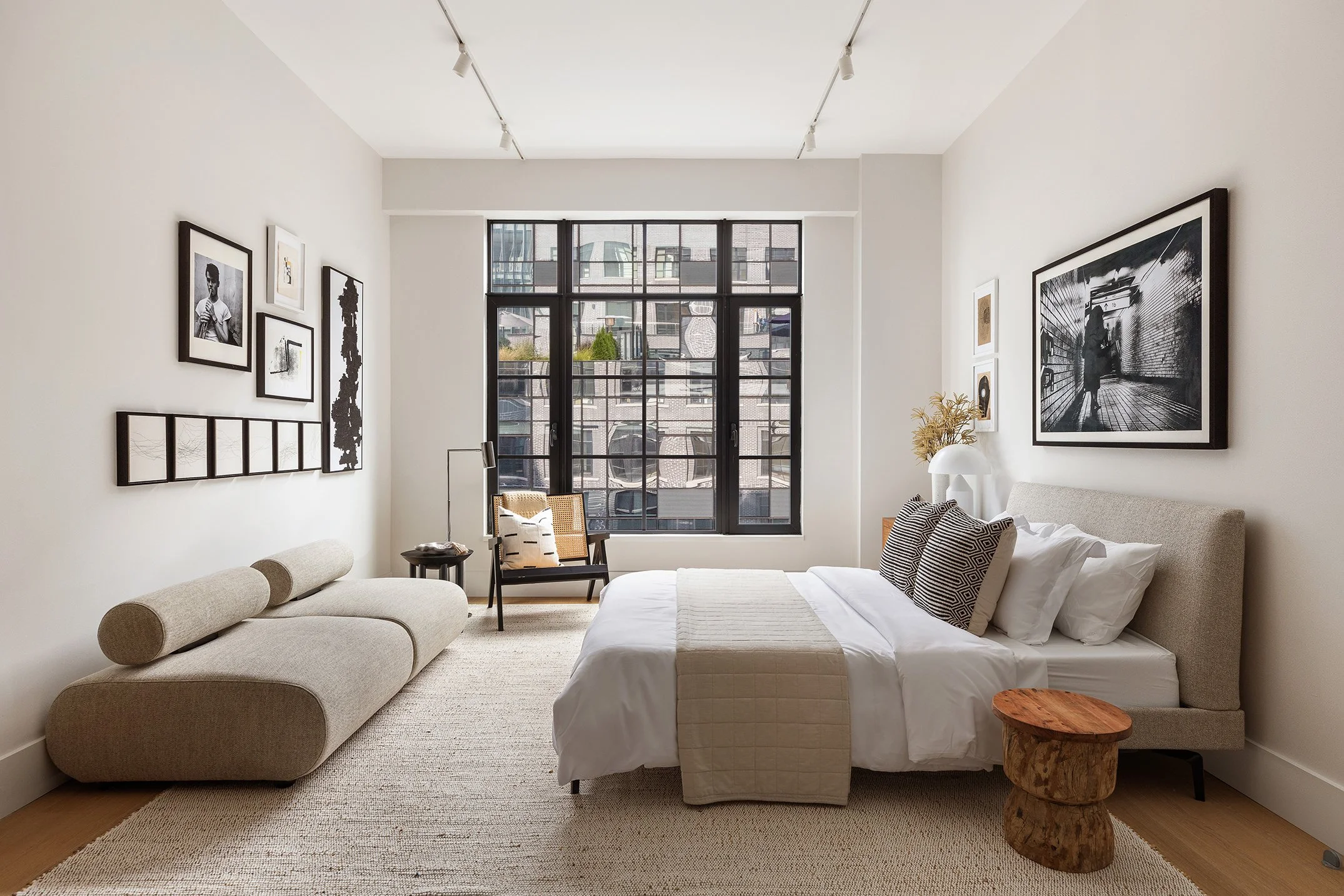 Art direction for staging - A modern, minimalist bedroom with large window, white walls, beige furniture, black and white artwork, and natural wood accents.