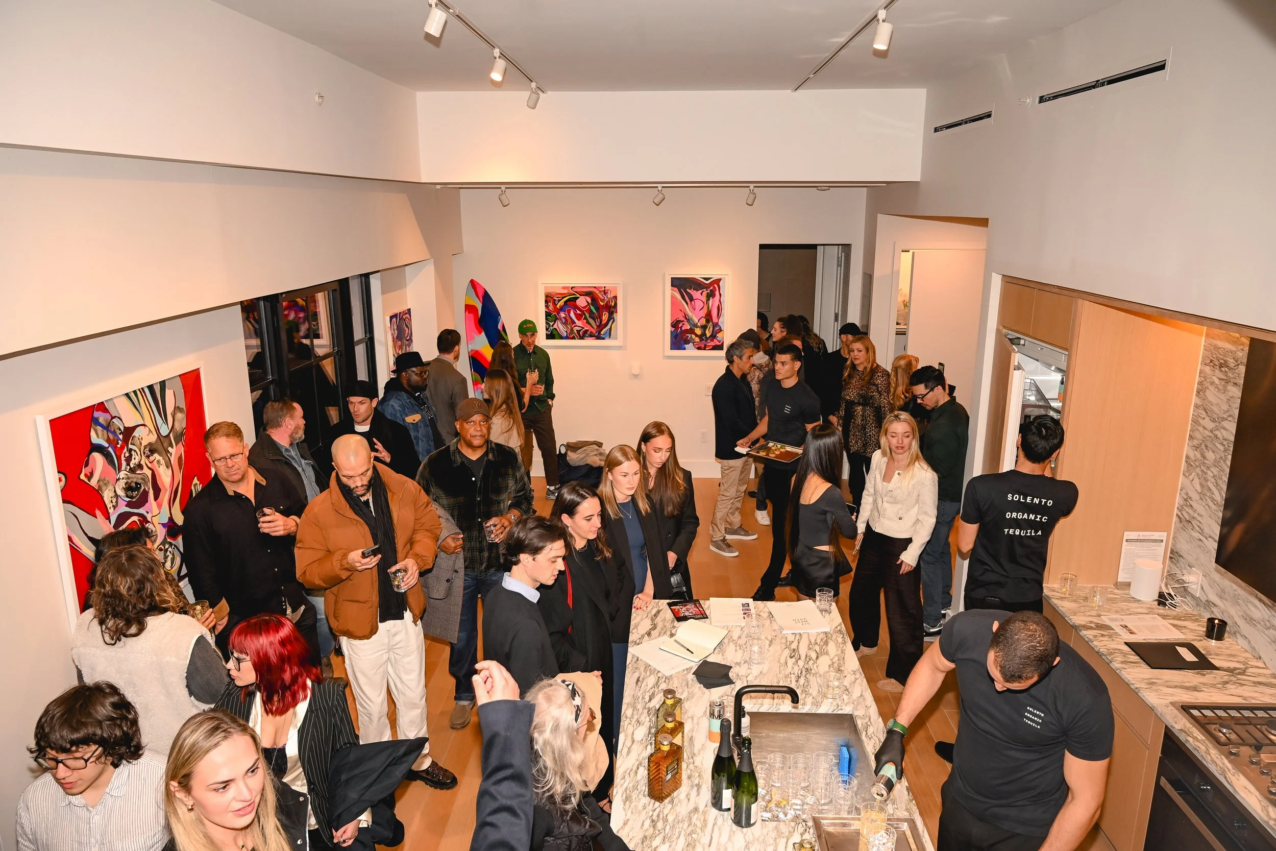 A crowded art gallery event with people mingling and viewing artwork. The walls feature colorful abstract paintings, and there is a bar area with bottles on a marble counter. Guests are dressed in various stylish outfits, and some are holding drinks.