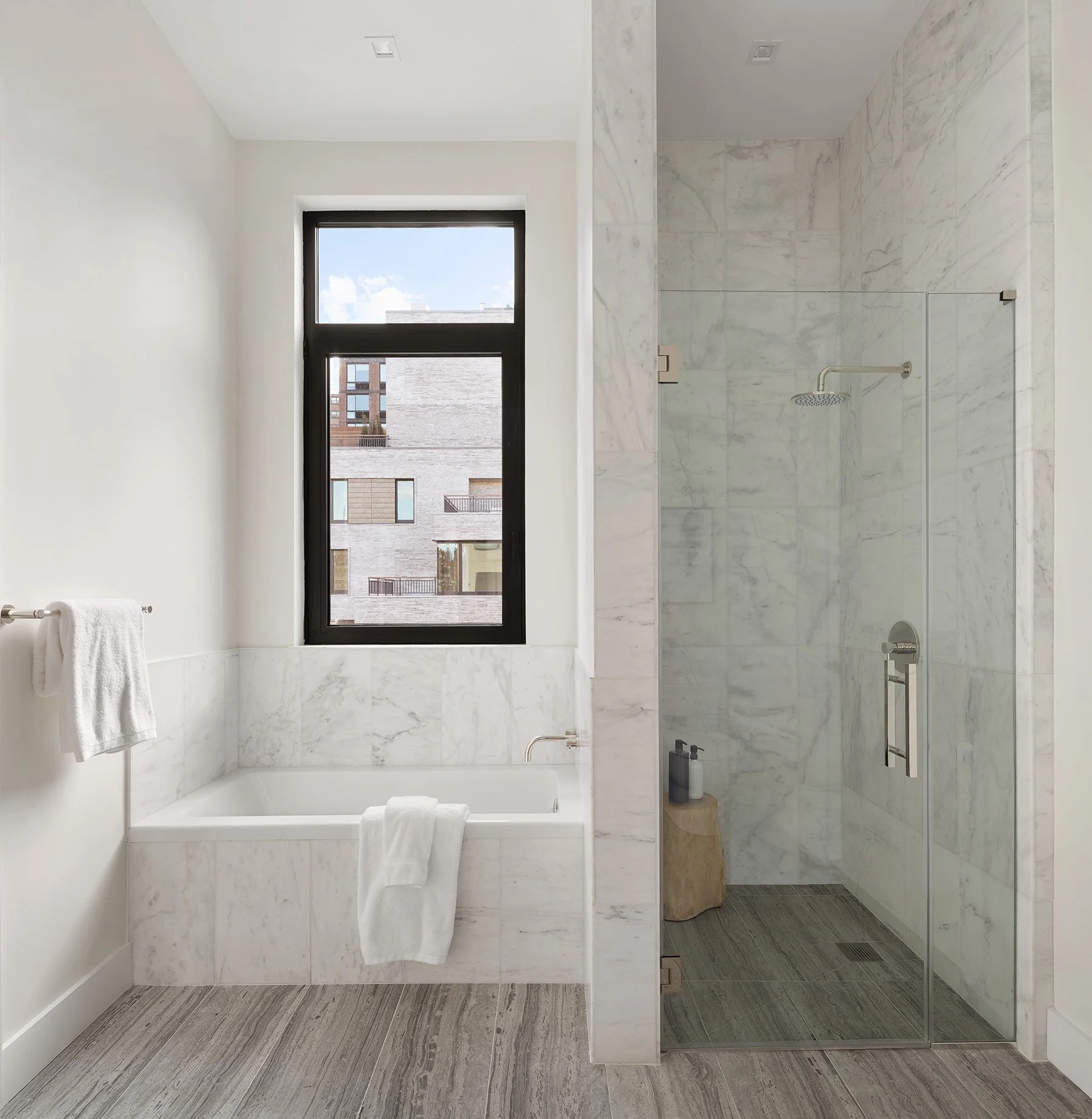 Staging Art direction - Bathroom with a bathtub and a large window on the left side, and a walk-in shower with marble walls and a glass door on the right side. The floor has gray wood-like tiles.