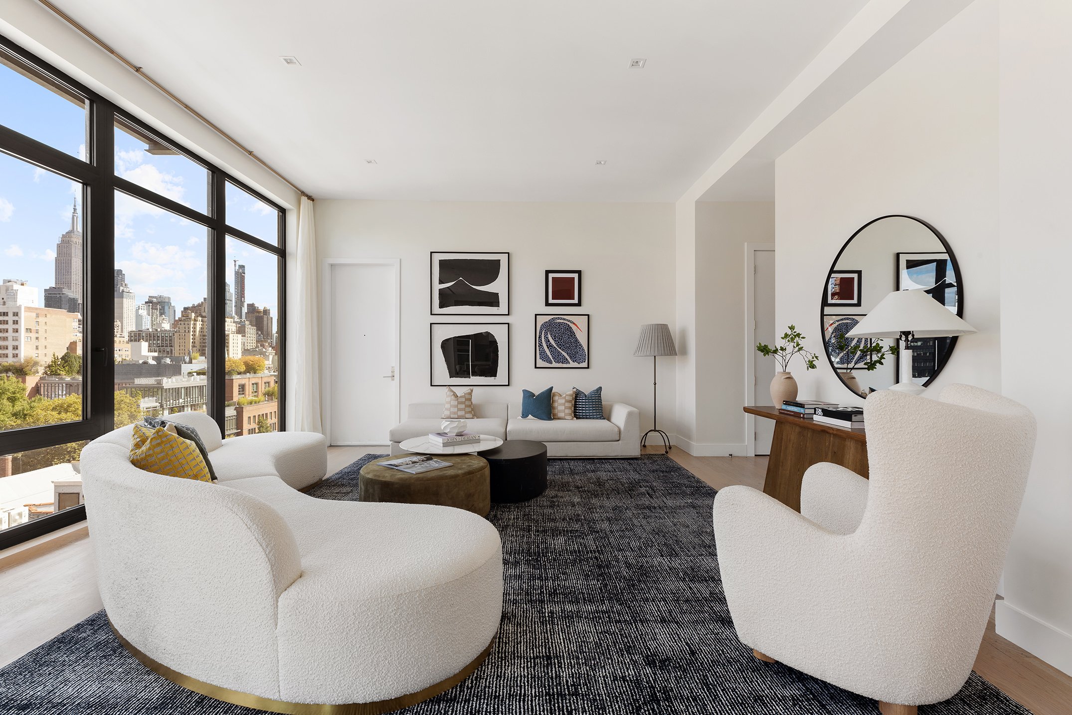 Staging Art direction - Modern living room with large floor-to-ceiling windows showing city buildings, white sofa with patterned cushions, abstract art on the wall,  floor lamp, and coffee table with magazines, all in a bright, minimalist style.