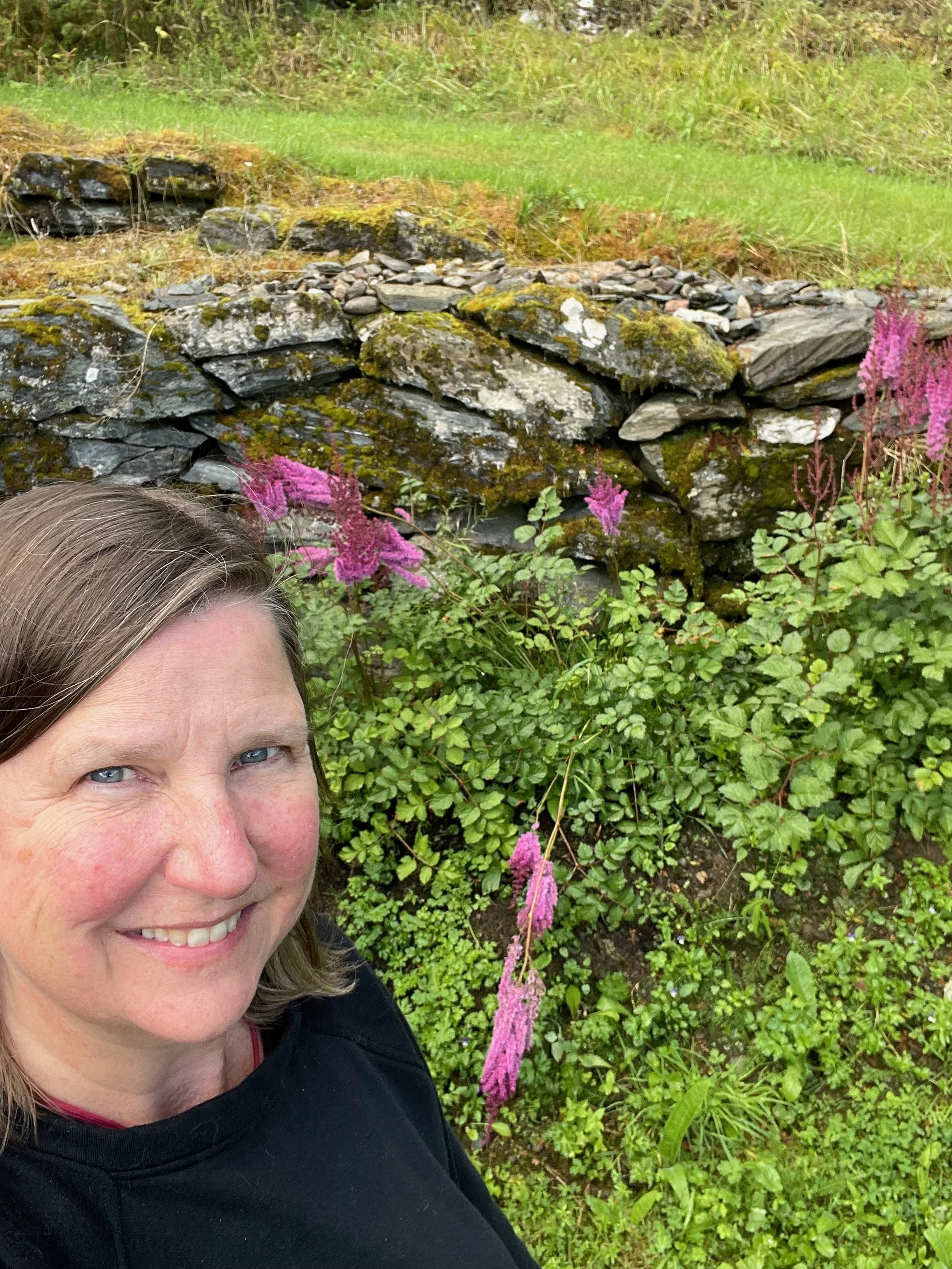A woman smiling outdoors in front of a moss-covered stone wall with pink wildflowers.