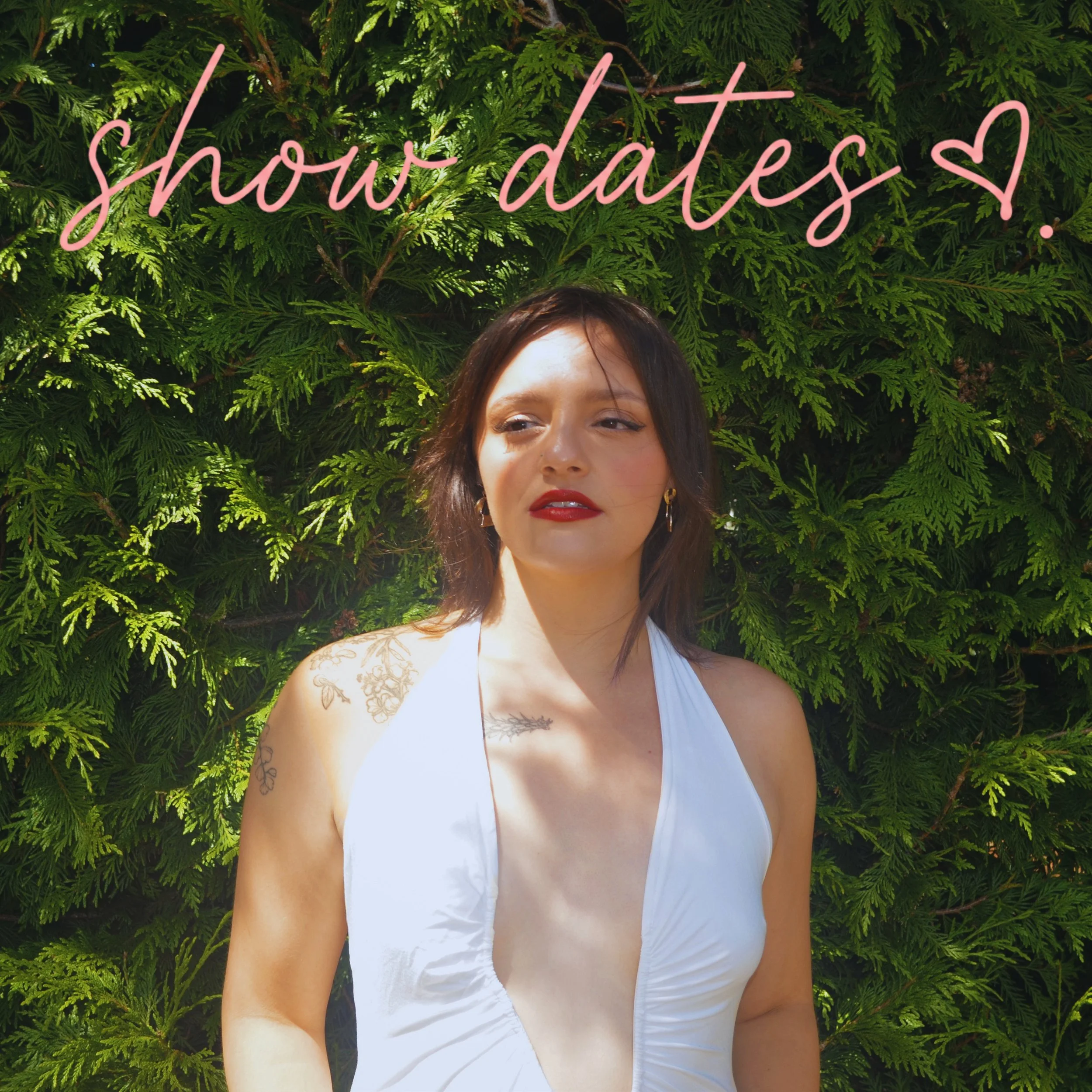 Young woman with dark hair and red lipstick wearing a white top, standing in front of lush green foliage with a pink neon sign that says 'shout outs' and a small heart.