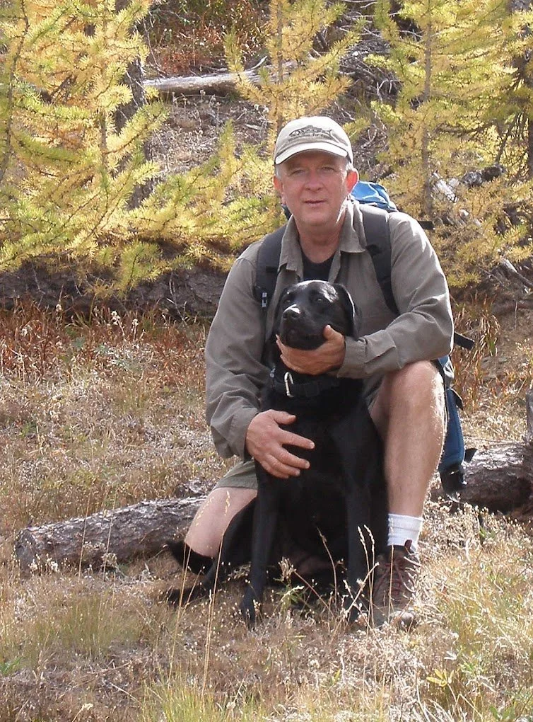 Acupuncturist Jim Dowling kneels outdoors on forest floor holding a black Labrador with a joyful expression, surrounded by yellow-green trees in the Methow Valley.