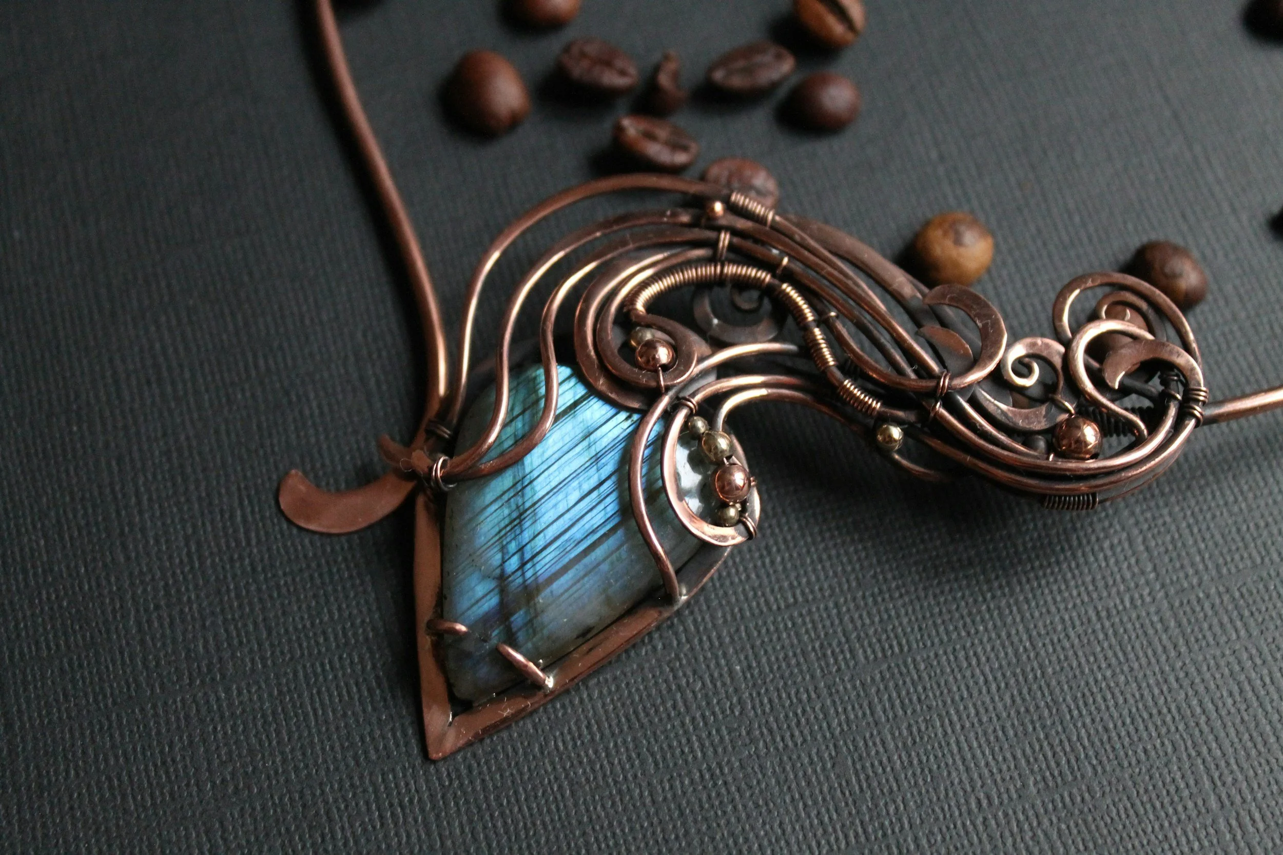 Close-up of a decorative necklace with a large blue stone pendant, surrounded by copper wire swirls, and coffee beans scattered on a dark fabric background.