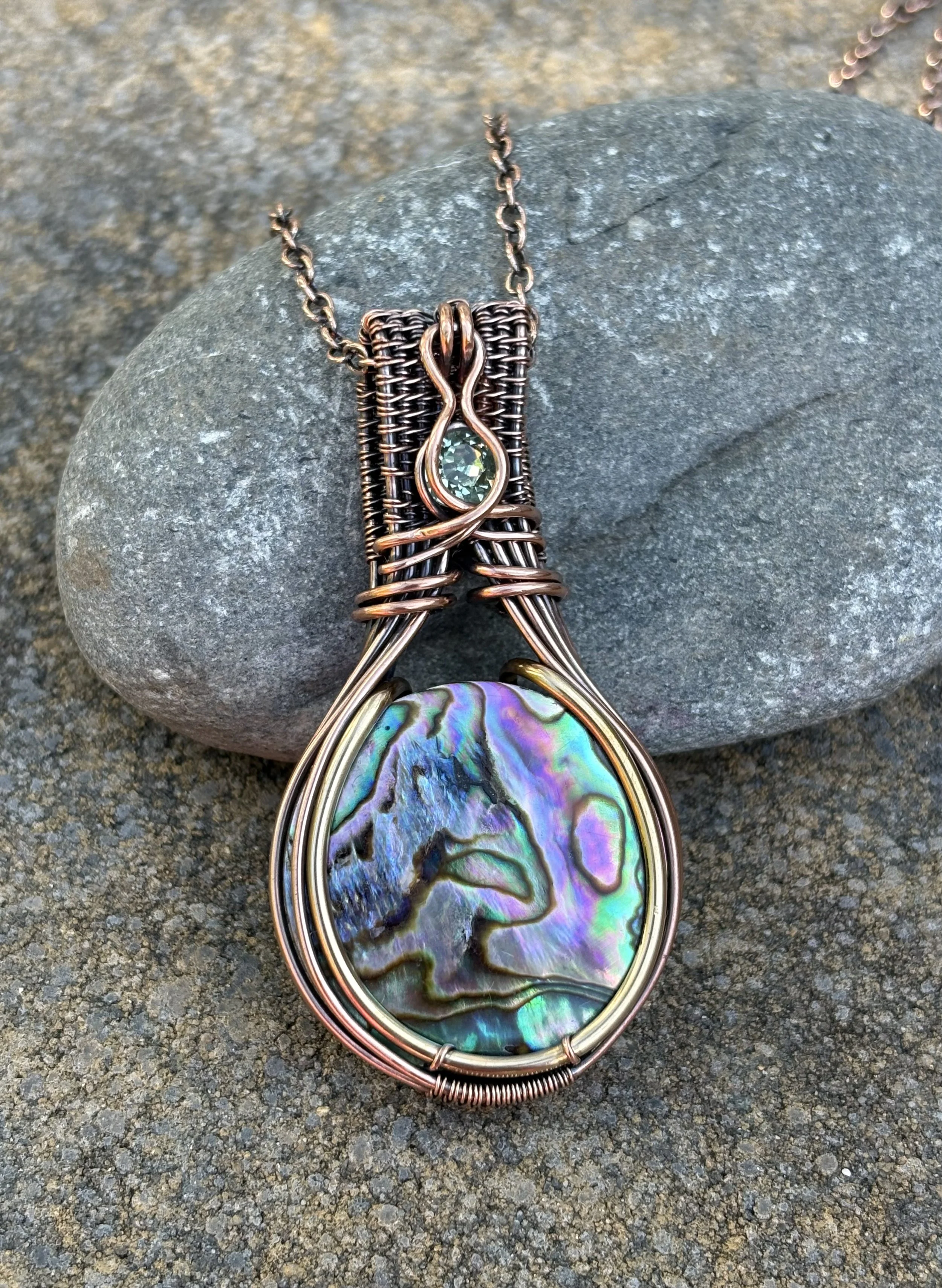 Close-up of a wire-wrapped pendant with a colorful, swirling abalone shell, hanging from a chain on a rough, gray stone surface.