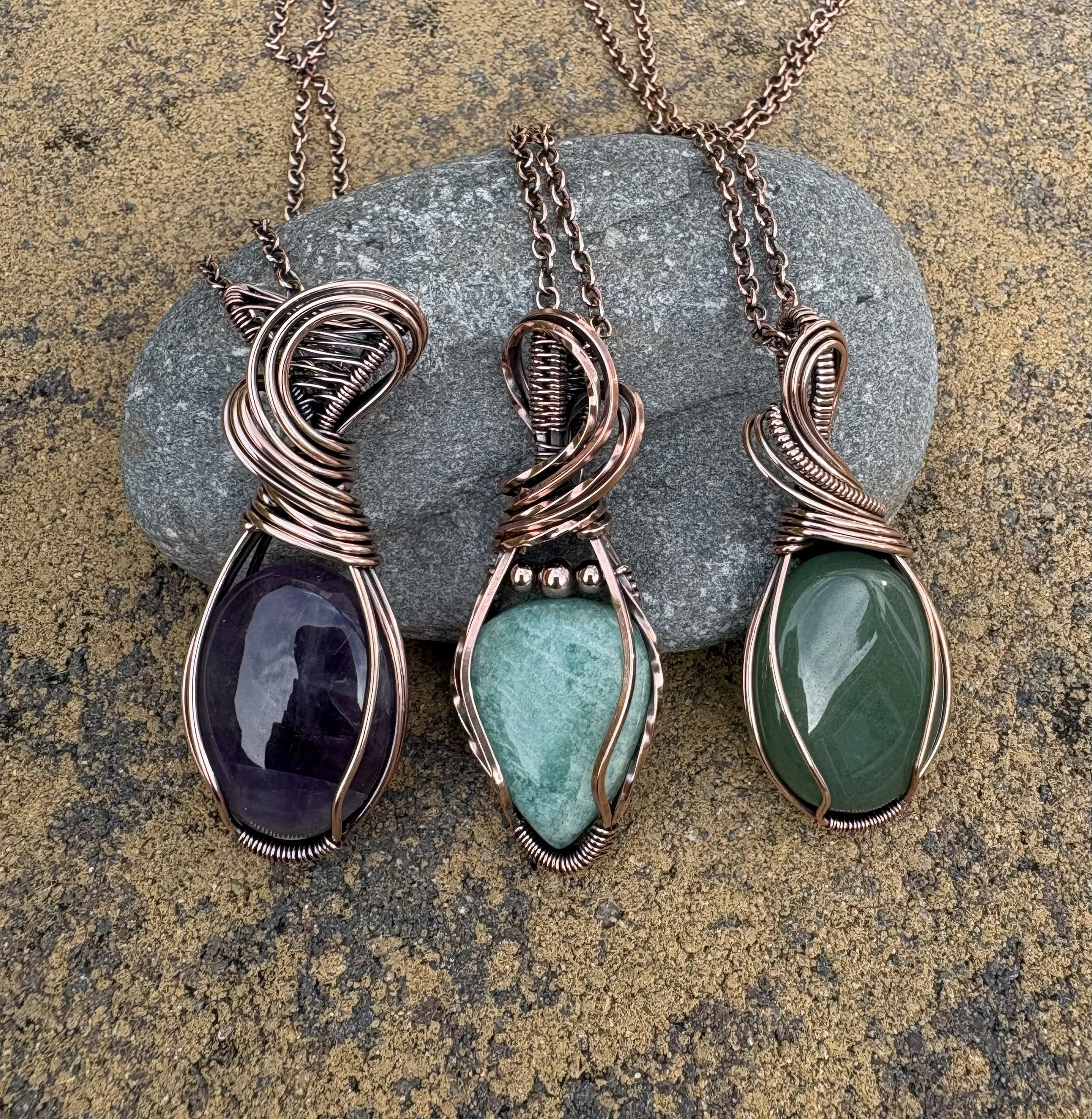 A collection of copper and green gemstone jewelry, including necklaces and earrings, displayed on a large rock on a gravel surface.
