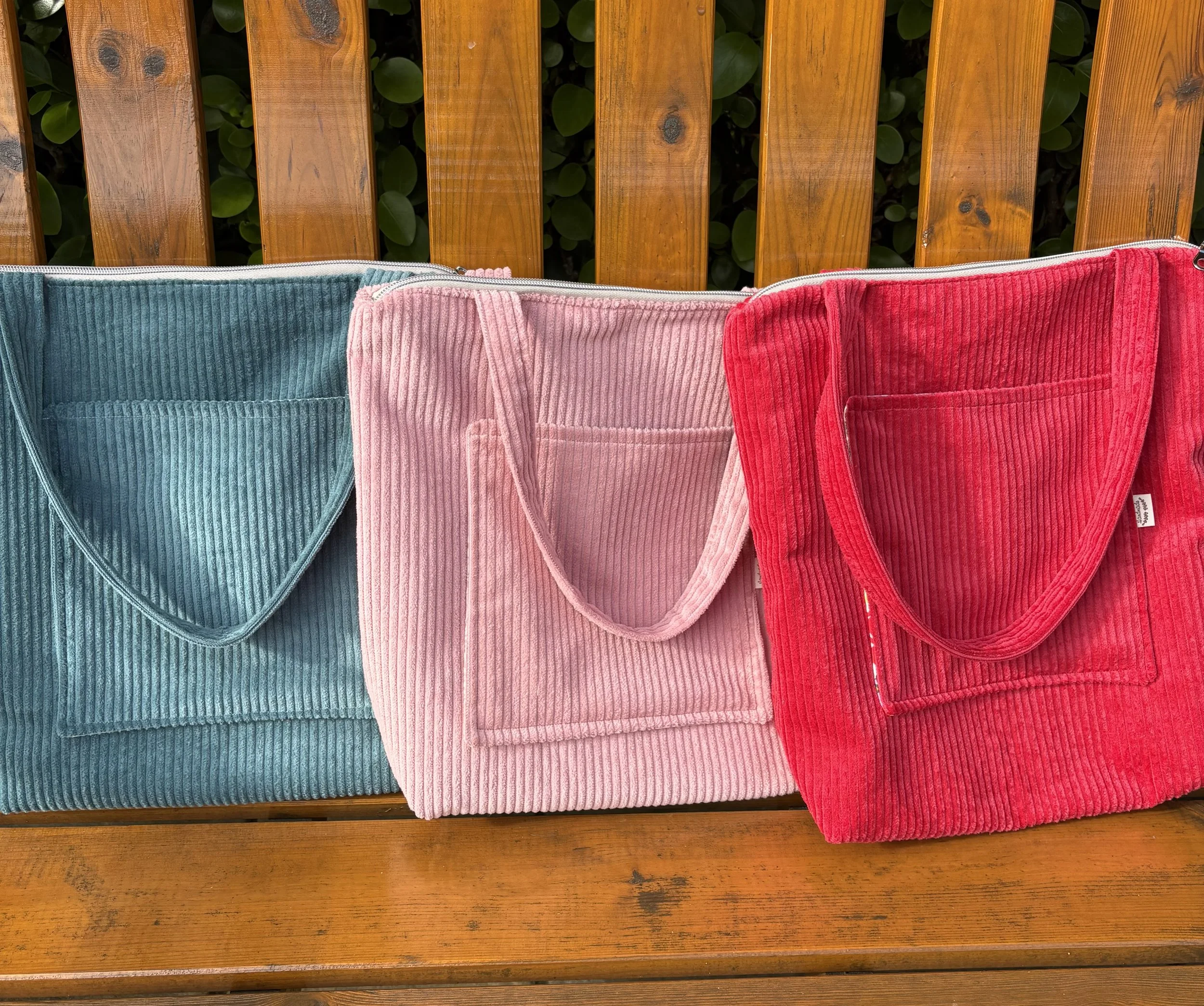 A row of three corduroy bags in teal, pink, and red hanging on a wooden bench with a background of green leaves.