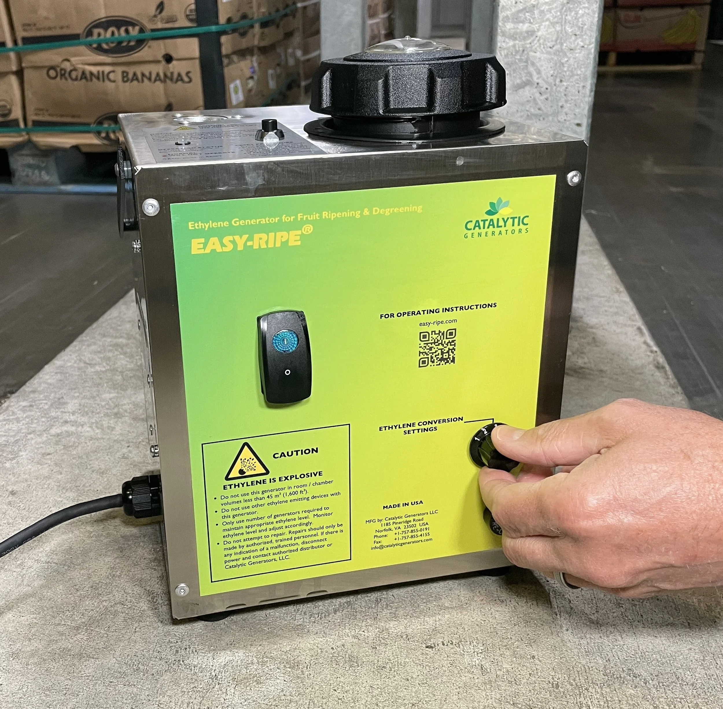 A person adjusting the settings on an Easy-Ripe ethylene generator for fruit ripening and degreening. The device is from Catalytic Generators and has a warning label about ethylene being explosive. The generator has a switch, QR code, and knob for ethylene conversion settings. Boxes of organic bananas are visible in the background.