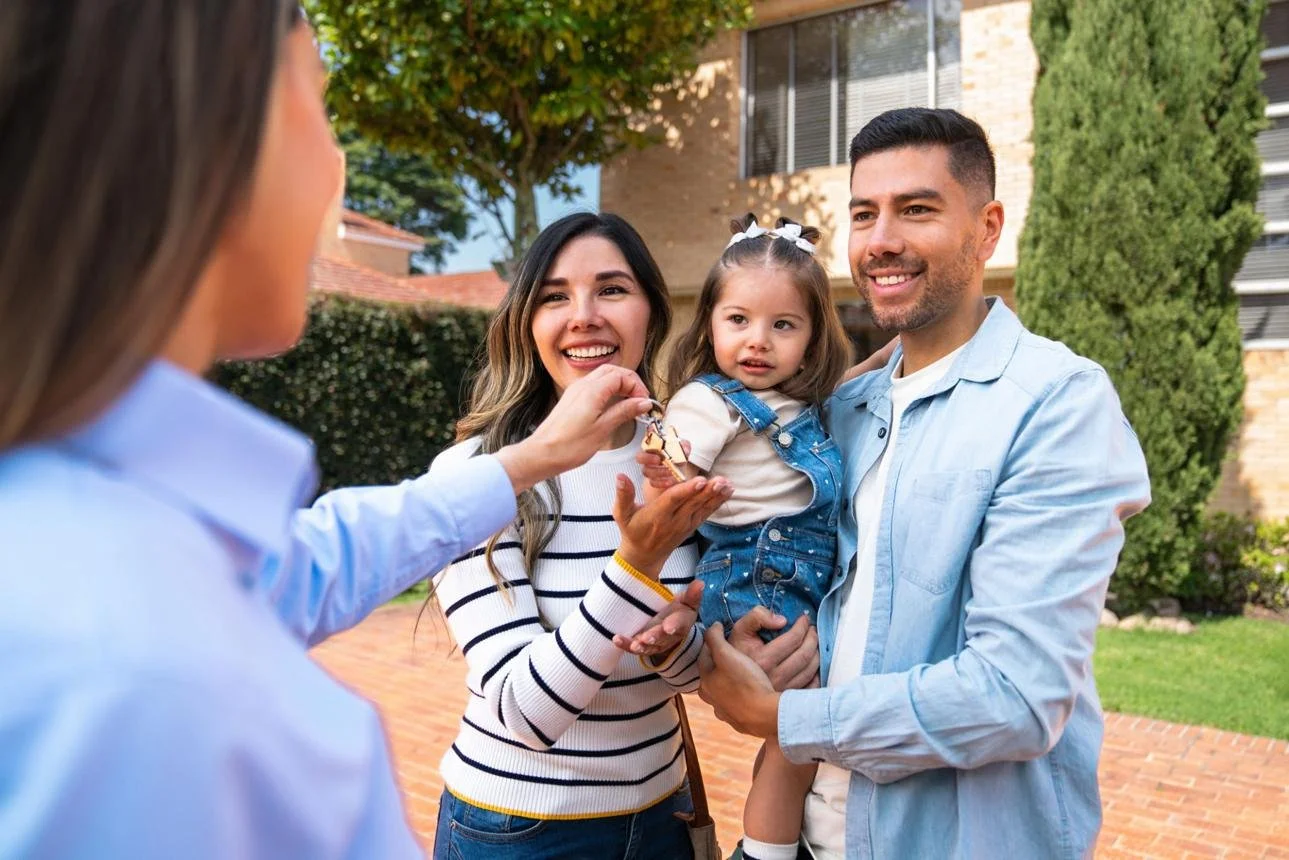 A family of three with a woman, a man holding a young girl, enjoying an outdoor gathering as a woman hands a small item to the girl during daytime.