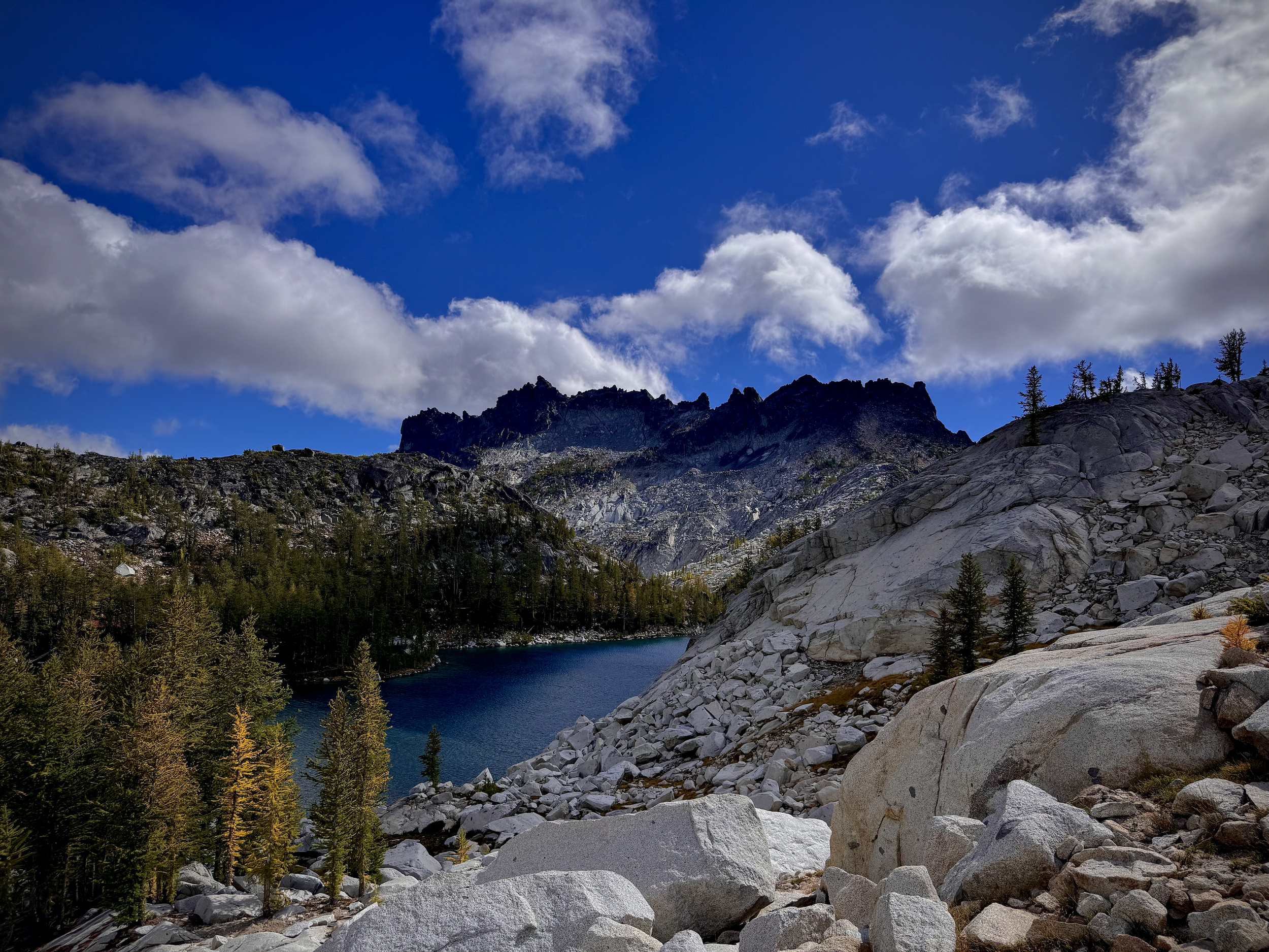 A mountain landscape with rocky slopes, a forested area, a lake, and a partly cloudy blue sky.