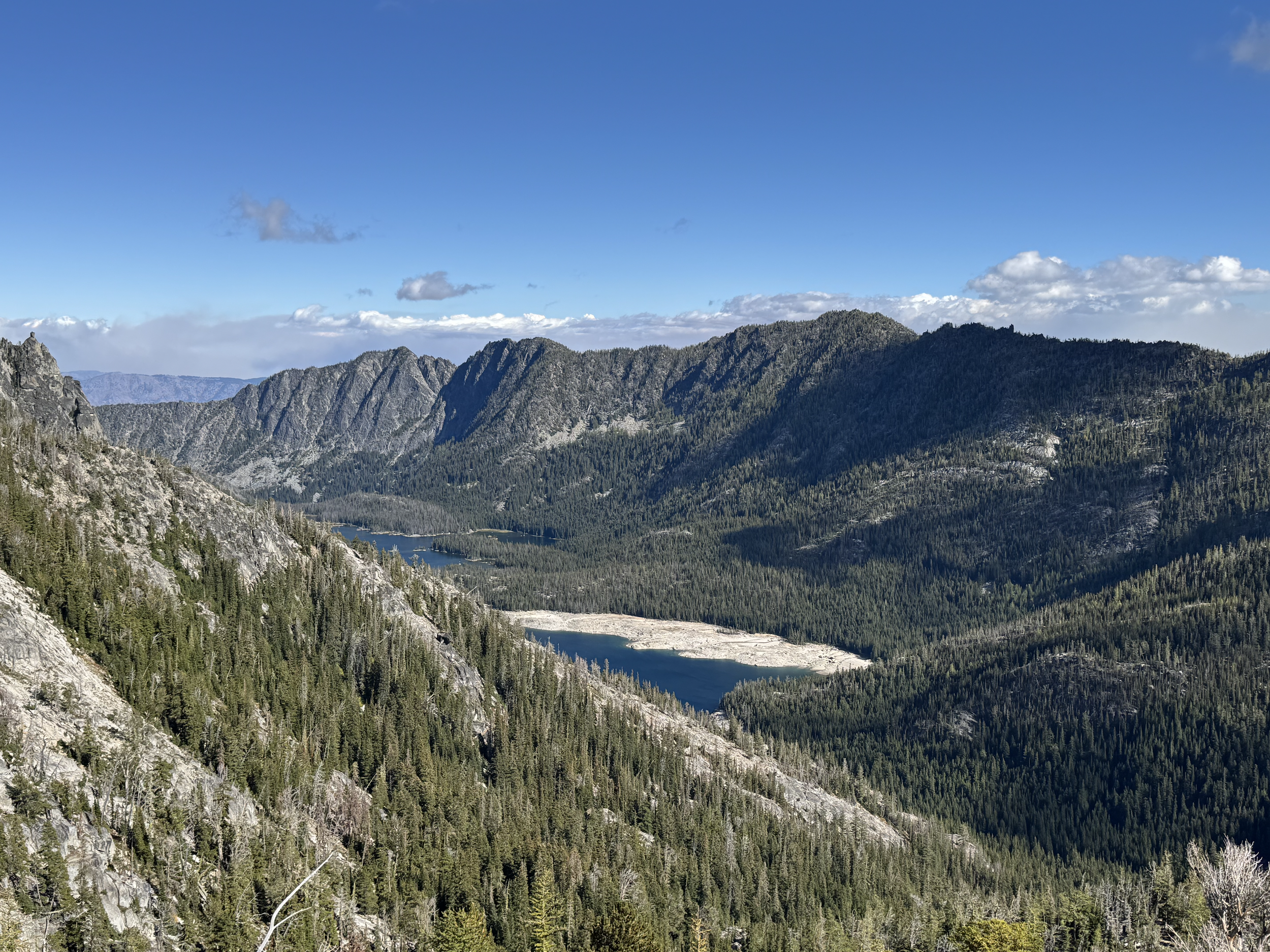 A panoramic view of a mountain range with dense forested slopes, a river or lake in the valley, and a partly cloudy sky overhead.