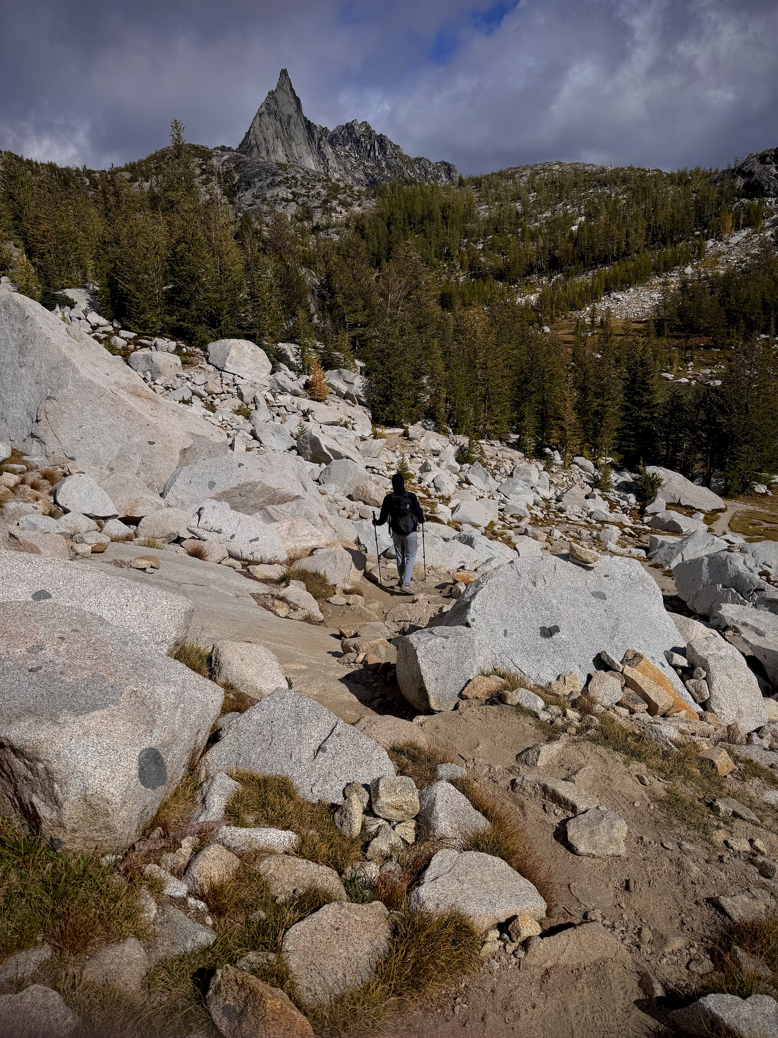 A person with a backpack and hiking poles walking on a rocky trail through a mountainous area, with tall trees and a prominent mountain peak in the background under a cloudy sky.