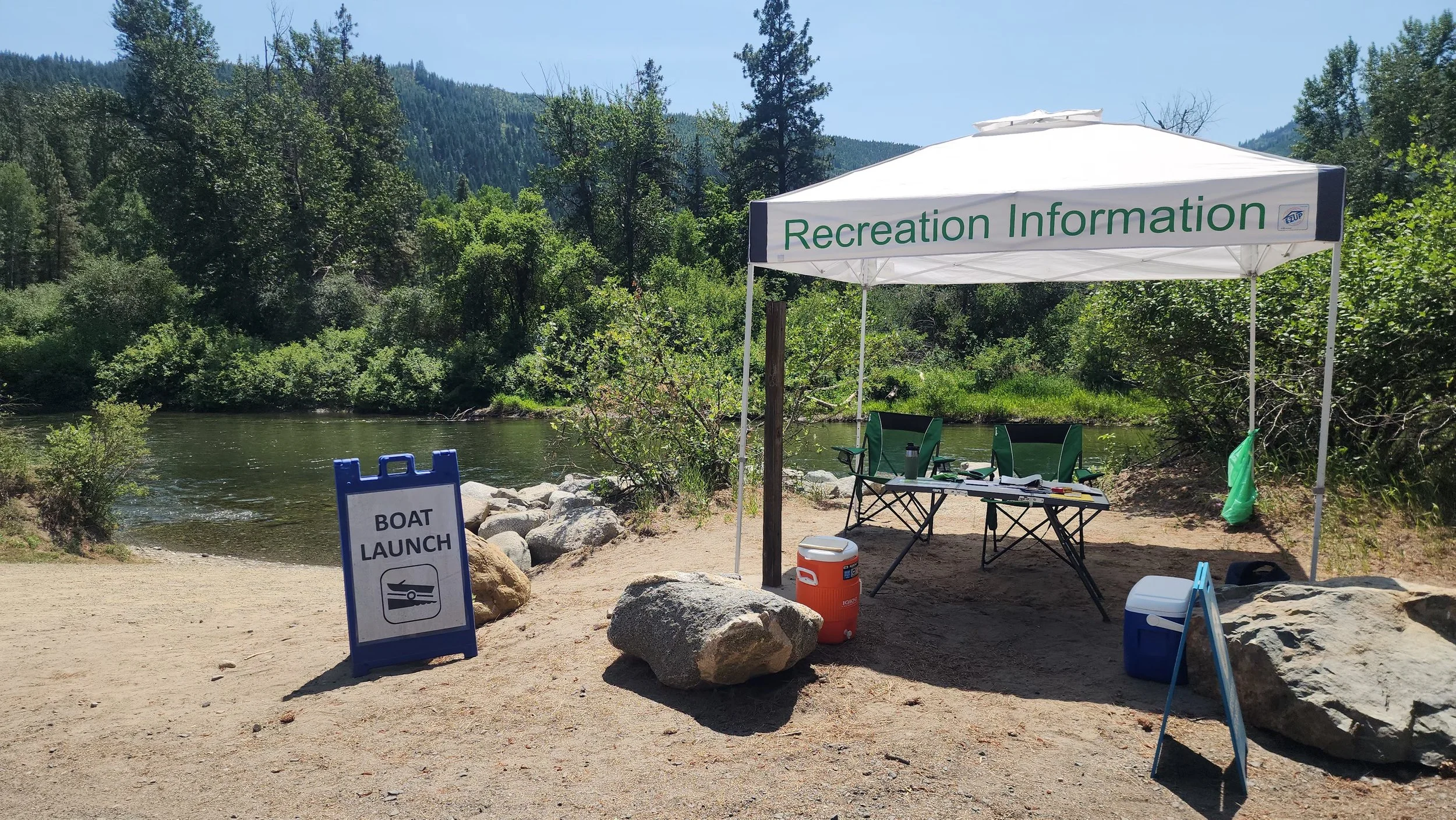 A recreation area by a river with a white tent labeled 'Recreation Information.' There are chairs, tables, and signs indicating boat launch and boat ramp. The surrounding area has trees and mountains in the background.